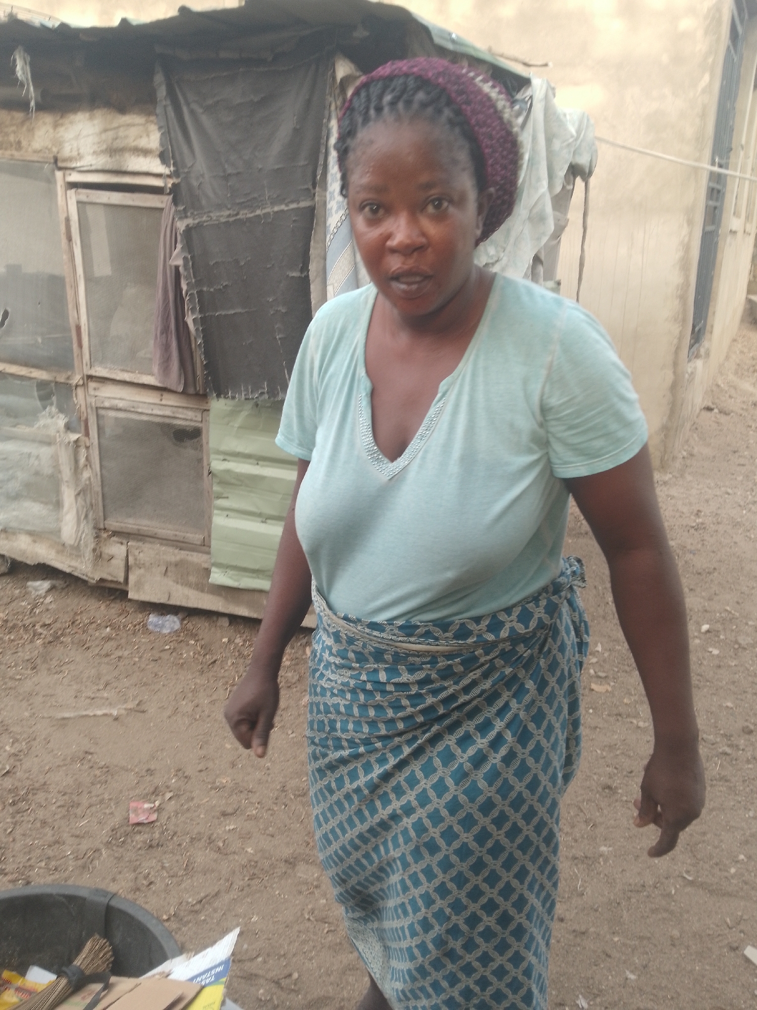 A woman stands in an outdoor dirt area in Lokoja, Nigeria. She has dark skin, is wearing a purple head covering, a light blue t-shirt, and a patterned blue and white wrap skirt. Behind her are two rundown structures: one appears to be made of patched-together materials, and the other is a building with a metal security grate over a window. To the left in the foreground is a dark gray plastic bin with refuse inside. The ground is dirt with scattered debris.