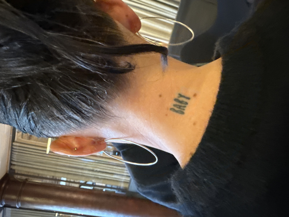 The scene depicts the back of an individual's head and neck, captured indoors within the city of London, United Kingdom. The subject has dark hair, styled back with some strands visible around the nape. Both ears feature large, silver-colored, oval hoop earrings. The left ear also has a small gold-colored stud piercing in its upper cartilage. A dark tattoo spelling "BABY" in uppercase letters is centrally located on the back of the neck, below the hairline. Several small moles are present on the neck skin. The individual is wearing a dark, possibly black, fuzzy-textured garment covering the upper back and shoulders. The background is out of focus, revealing a reddish-brown fluted column on the left and light-colored vertical blinds or drapes further back.