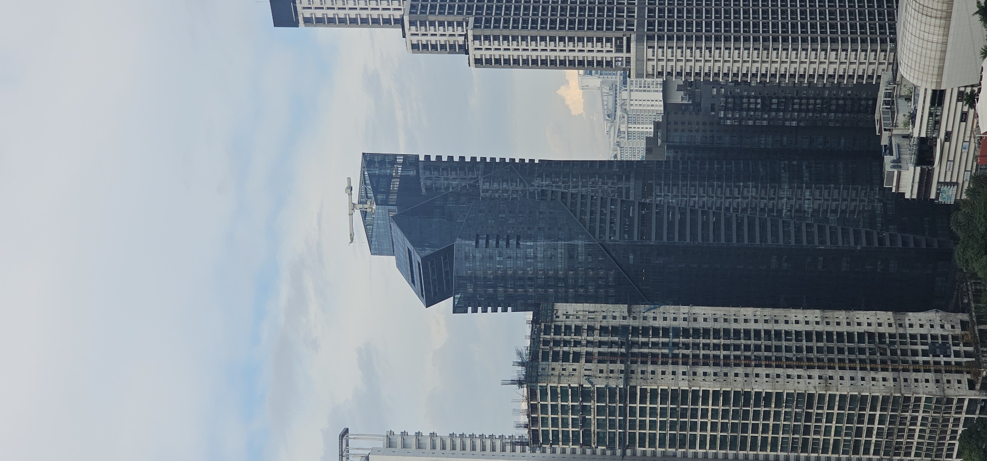 The image depicts a cityscape of Makati, Philippines, dominated by tall buildings under a cloudy sky. In the center, a striking, dark-blue skyscraper with a distinctive angled design stands out. A construction crane is visible on its rooftop, suggesting ongoing development or perhaps an antenna. To the left, a tall, white building is partially visible, appearing to be under construction with exposed concrete and rebar. To the right and in the background, several other high-rise buildings of varying designs and construction stages fill the frame. The lower part of the image shows more buildings, some appearing to be completed commercial structures with visible signage and some green foliage. The overall scene conveys a sense of urban development and a bustling metropolitan environment.