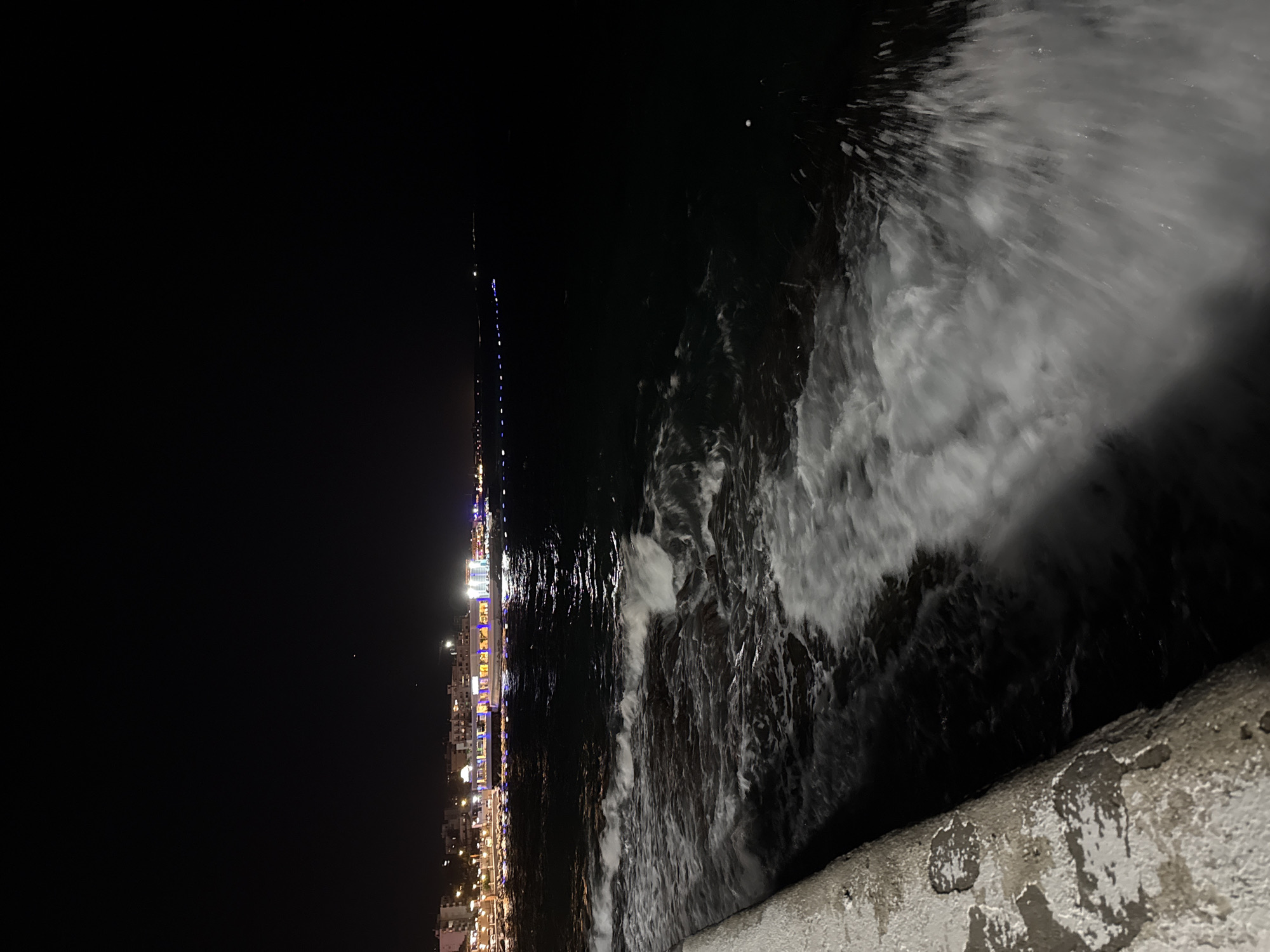 The image is a nighttime view of a coastline with waves crashing against a concrete barrier in the foreground. In the distance, a city skyline is illuminated by numerous lights, suggesting a vibrant urban area. The sea is dark, with the white foam of the waves providing a stark contrast against the black water. A few faint lights are visible on the horizon, possibly indicating ships or other structures in the distance. The overall atmosphere is one of tranquility and a touch of mystery due to the darkness and the crashing waves.