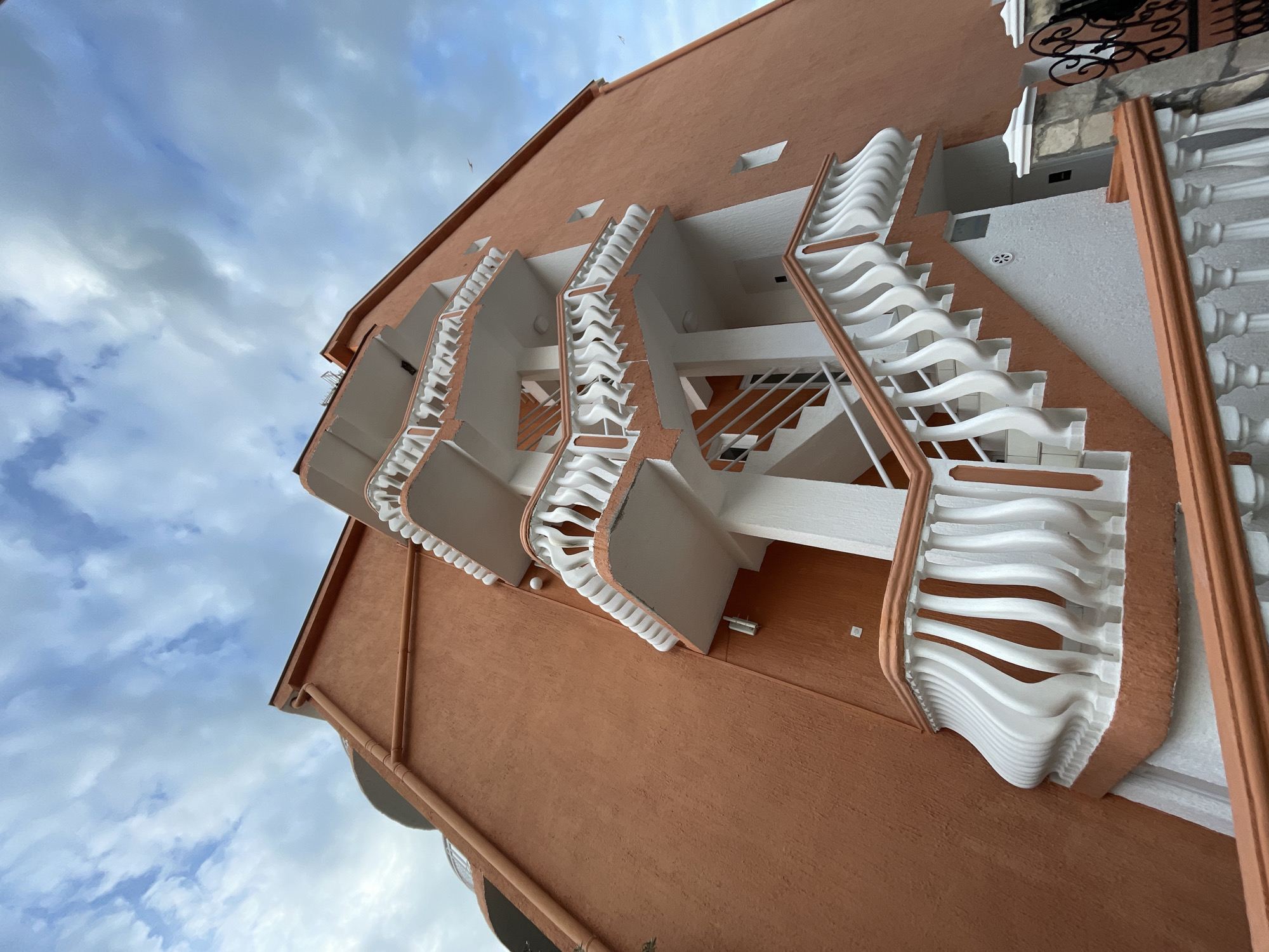 An upward-angled view of a multi-story building in Rafailovići, Montenegro. The building's exterior is predominantly orange, with white balconies featuring distinctive curved railings. Staircases are visible within the balcony structures. The sky in the background is blue with scattered white clouds.