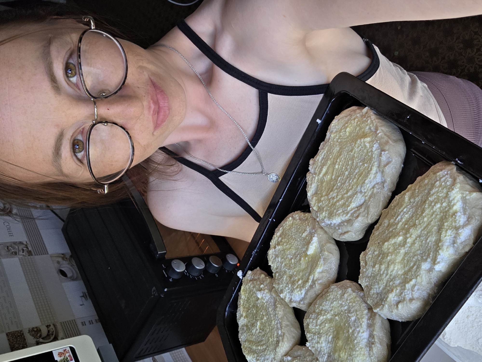 A woman wearing glasses and a beige tank top with black trim holds a baking tray with six circular, uncooked dough portions topped with a light-colored, creamy substance. The dough appears to be either bread or pastry. Behind her, a black electric oven is visible with its door ajar and the interior light on. The scene appears to be set in a kitchen, likely in an apartment within the city of Chaikovsky, Russia, given the context of the request. The lighting suggests daytime, and the overall environment is domestic.