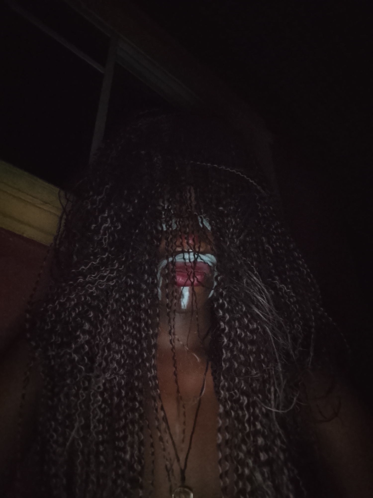 A person with long, dark, curly hair covering most of their face is visible in a dimly lit environment. The face that is visible has white markings across the upper lip and chin. A pendant hangs around their neck. The setting appears to be indoors. The city and specific location are unknown.