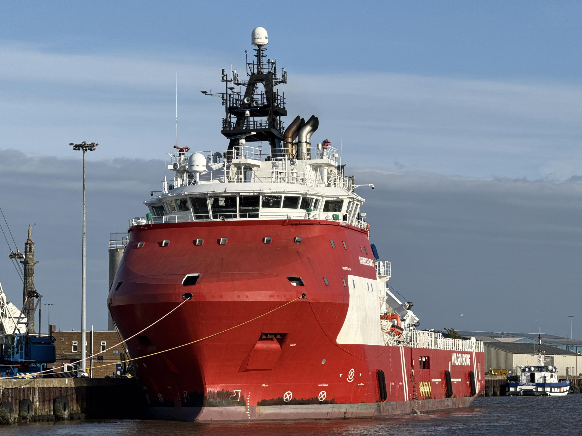 The image shows a large, red offshore supply vessel, named "Keizersborg" and "Wagenborg", docked at a port in Gorleston-on-Sea, United Kingdom. The vessel is tied to the dock with yellow mooring lines. The hull is primarily red with white sections and black rubber fenders along the waterline. The superstructure includes a white bridge with numerous windows, a black lattice tower with communication equipment, and exhaust stacks. In the background, there are buildings, a crane with a statue of Neptune on top, and a blue and white pilot boat. The sky is blue with some scattered clouds. A lamppost stands on the dock near the bow of the vessel. The water is calm.