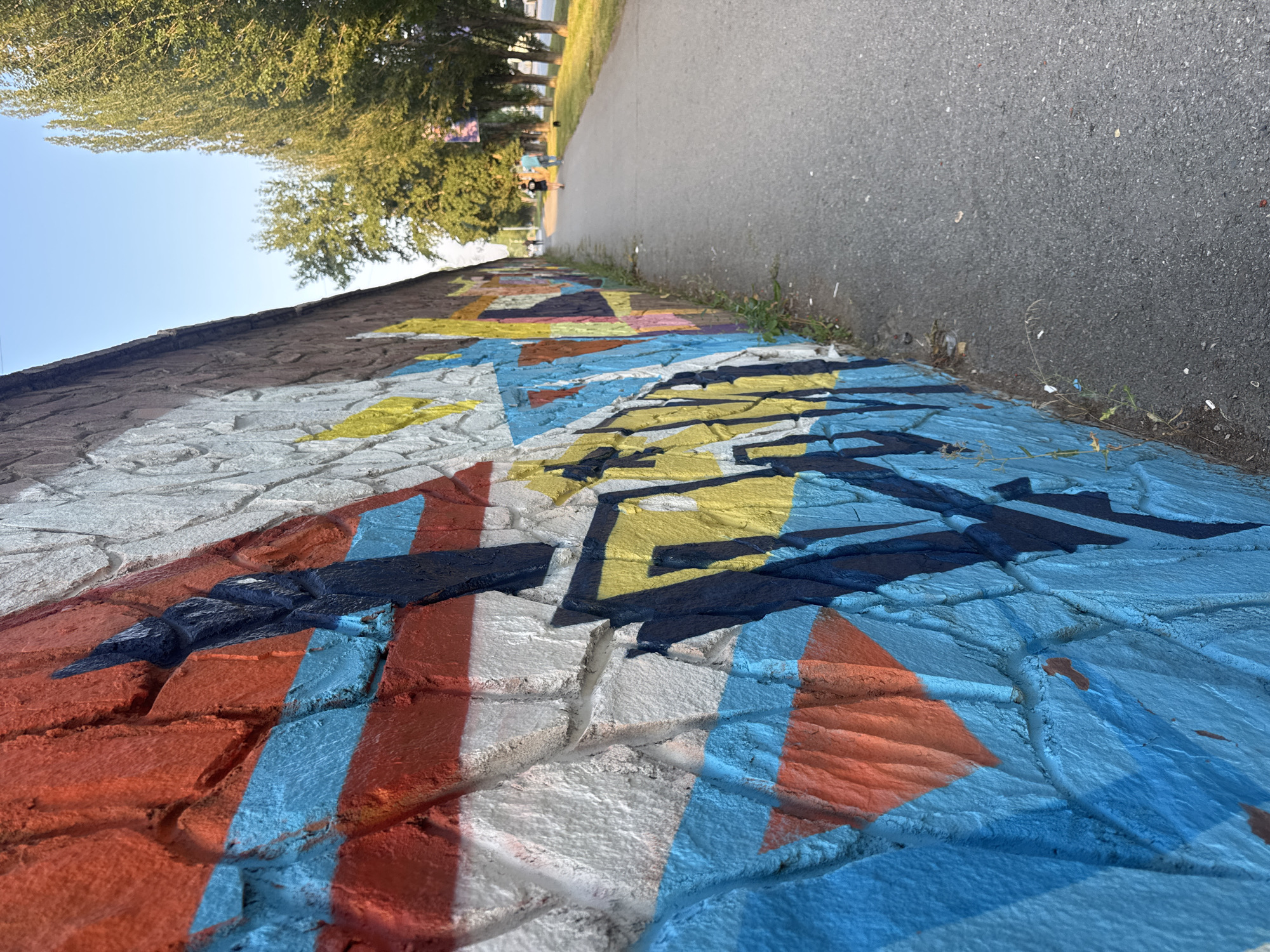 The image depicts a graffiti-covered stone wall alongside a paved walkway in Ufa, Russia. The wall is painted with a vibrant abstract design featuring bold strokes of red, orange, blue, yellow, and white. Dark blue and black paint is used for outlines and angular shapes. The walkway is a grey asphalt path that stretches into the distance, bordered by a grassy area and a line of tall, leafy trees. The sky above is clear and blue. In the background, several individuals are walking on the path, with one person appearing to be wearing a dark shirt and shorts, and another a light blue shirt and dark pants. A small, dark-colored dog is also visible on the walkway. The overall scene suggests an urban outdoor environment during daylight hours.