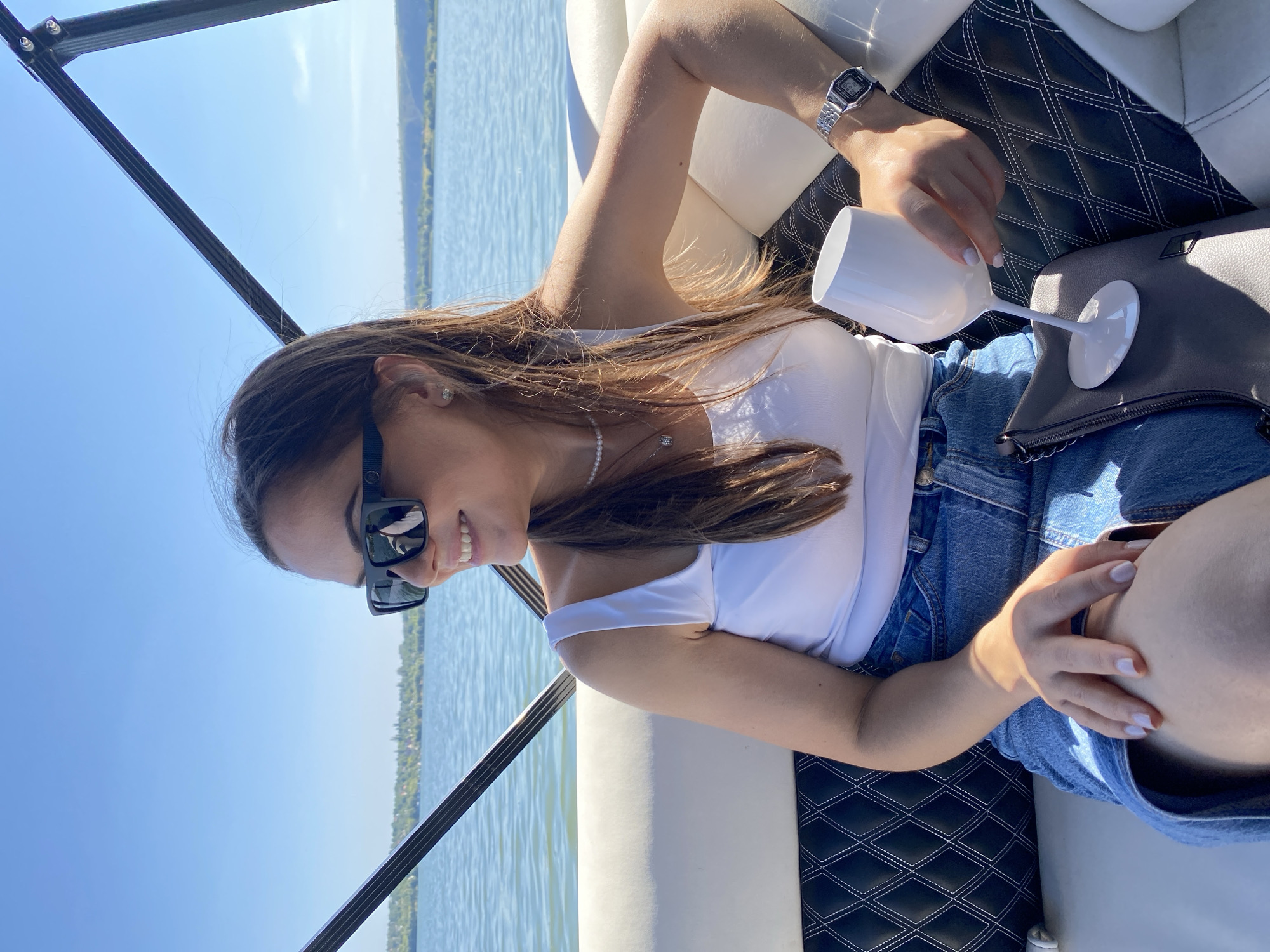 A woman with brown hair, wearing sunglasses, a white tank top, and a denim skirt, sits on a boat with a white and black upholstered interior. She is holding a white wine glass and her left hand rests on her lap. The boat is on a body of water, with a tree-lined shore in the background. The sky is clear and blue. The image appears to have been taken during the day. The location is the city of Molovata, Moldova.
