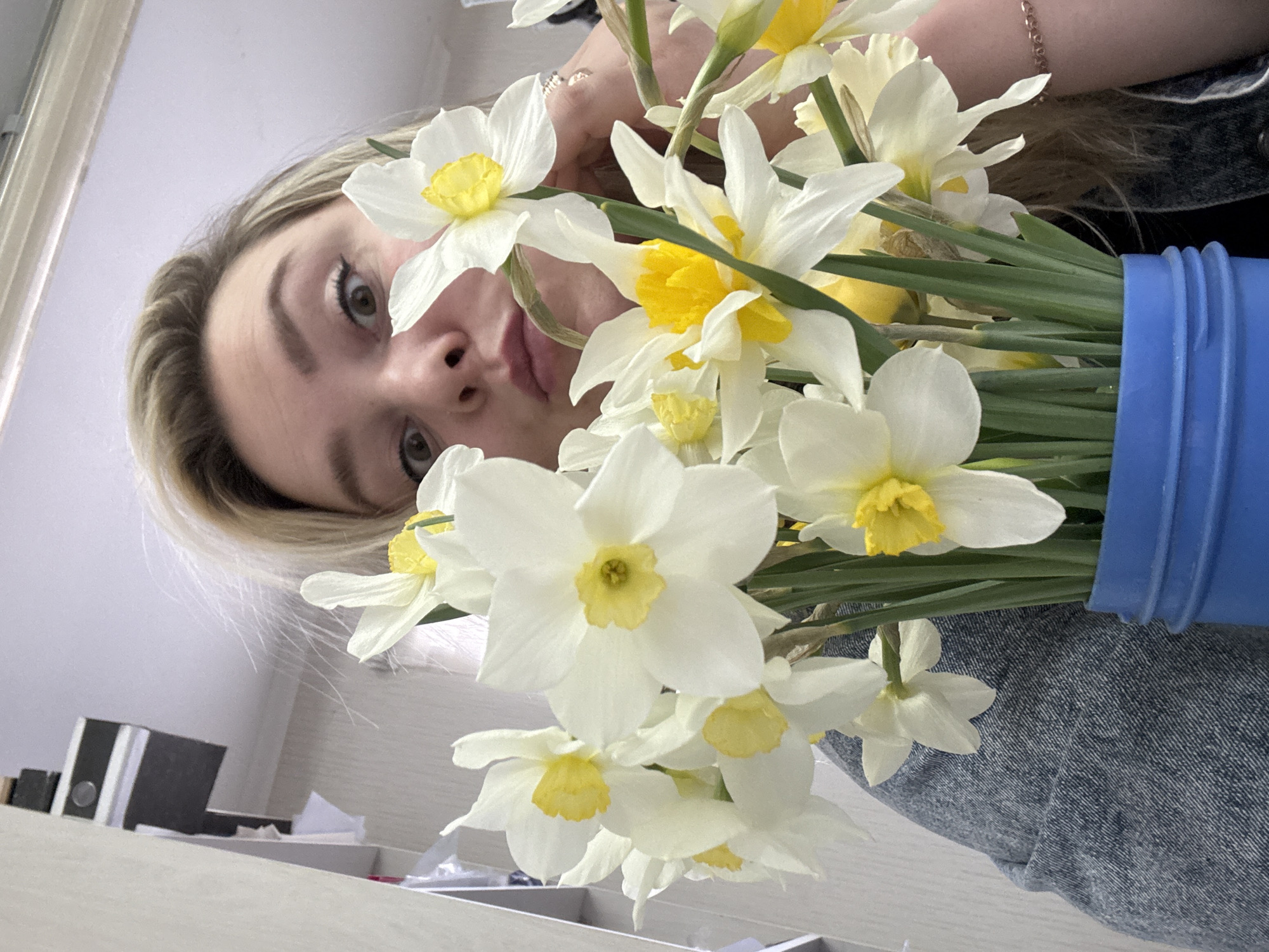 A woman with blonde hair is holding a bouquet of white and yellow daffodils in a blue container. She is wearing a denim jacket and a bracelet. The photo is taken from a low angle, looking up at the woman and the flowers. The background is a plain wall and a desk with office supplies. The location, Noviy, Russia, is not evident in the image.