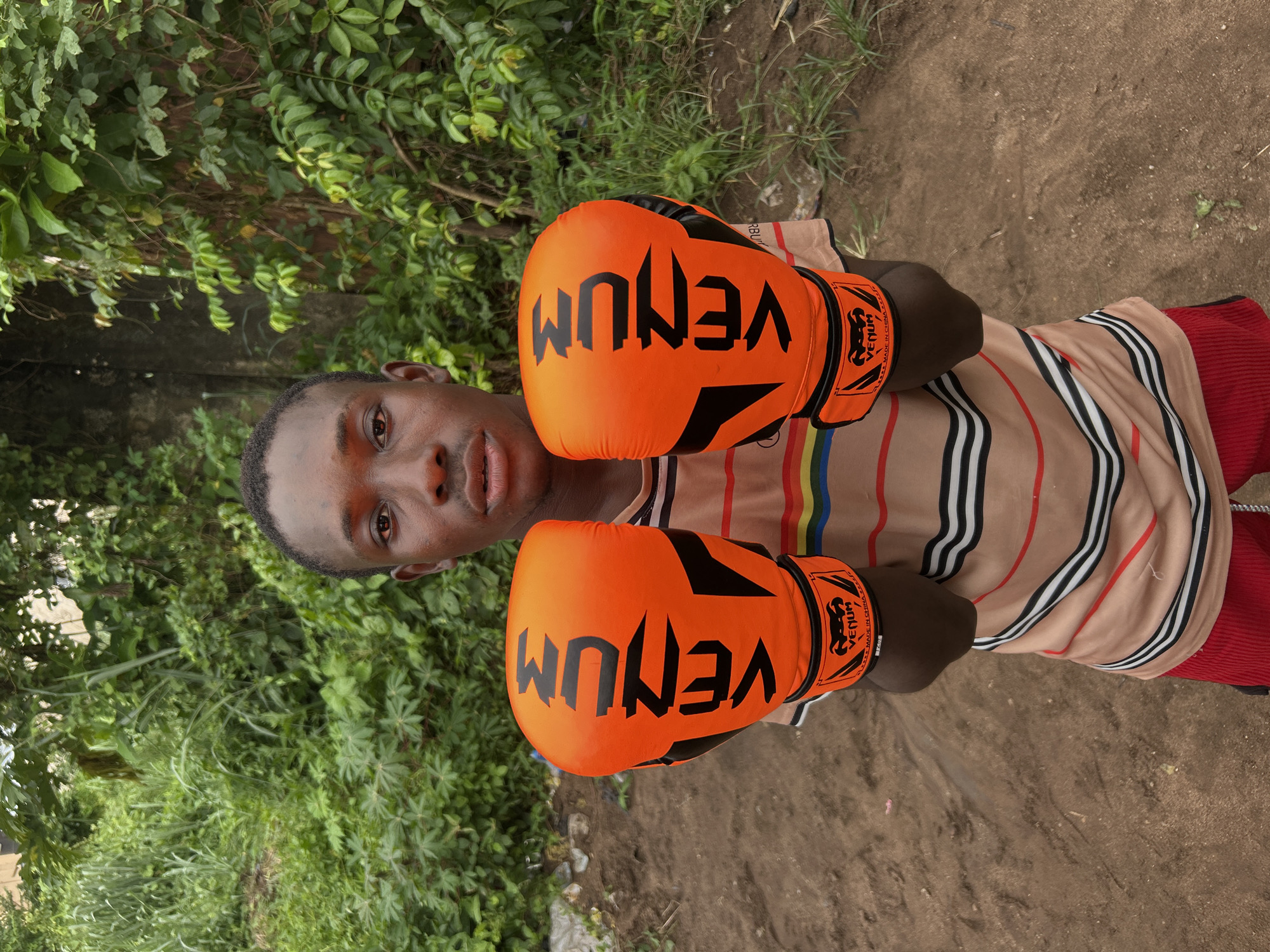 A young Black male wearing a striped t-shirt and red shorts stands outdoors in a dirt area, holding up two bright orange Venum boxing gloves. He is positioned centrally in the frame, facing the camera with a neutral expression. The background consists of lush green foliage and a wall. The scene appears to be in a residential area of Ihiagwa, Nigeria.
