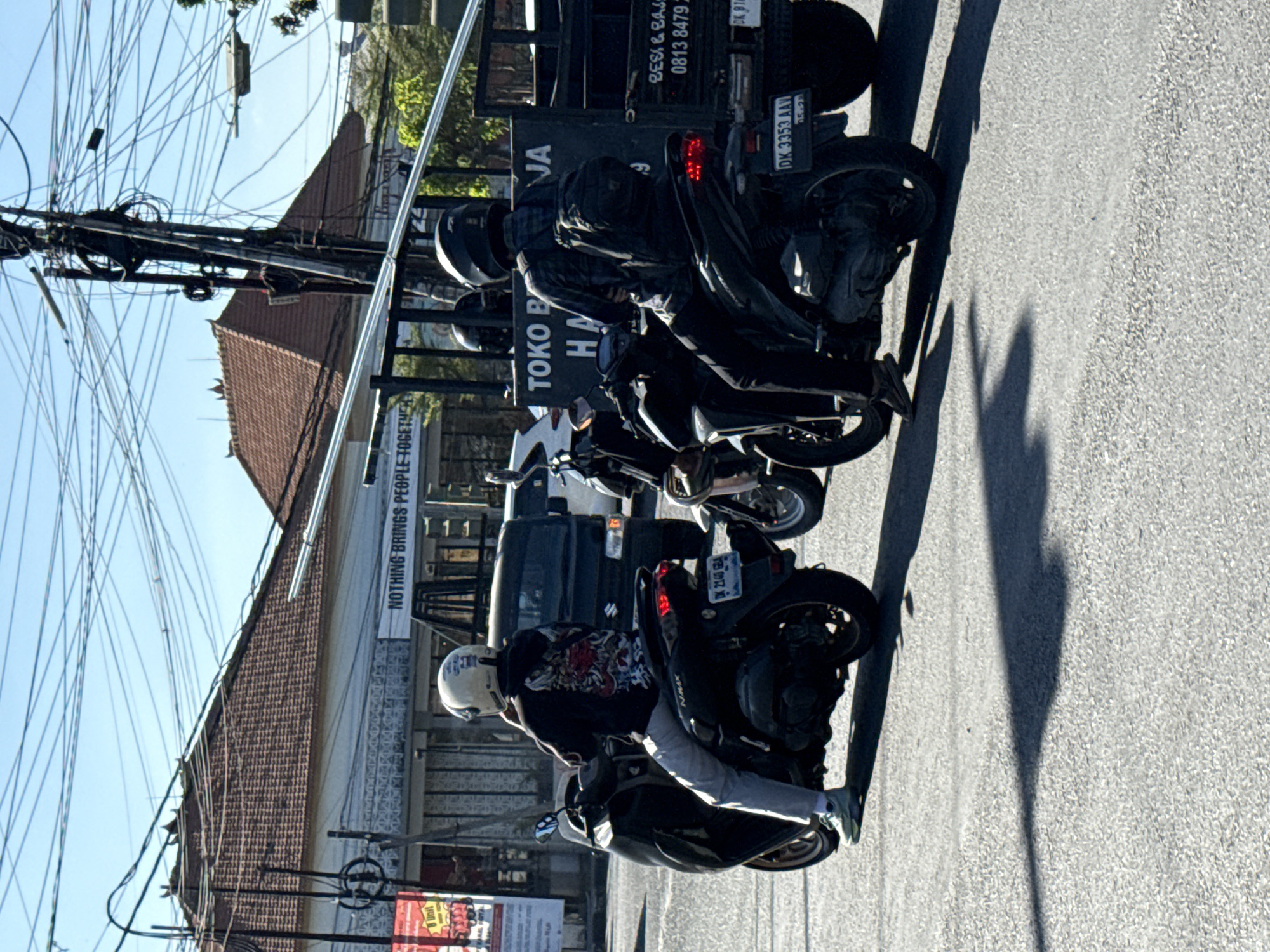 The image captures a street scene in Denpasar, Indonesia, under a clear blue sky. In the foreground, several motorbikes are visible, with riders in casual attire. One person on a black scooter is wearing a white helmet with graphics and a dark hoodie with an intricate design on the back. Another rider on a black motorcycle is wearing a dark helmet and a plaid shirt, with a backpack on. A dark-colored truck with signage is also present in the scene. The background reveals buildings with tiled roofs, a utility pole with numerous wires, and lush green trees. The street is paved, and shadows are cast on the road, indicating the time of day. The overall impression is a snapshot of daily life and transportation in a bustling urban environment.