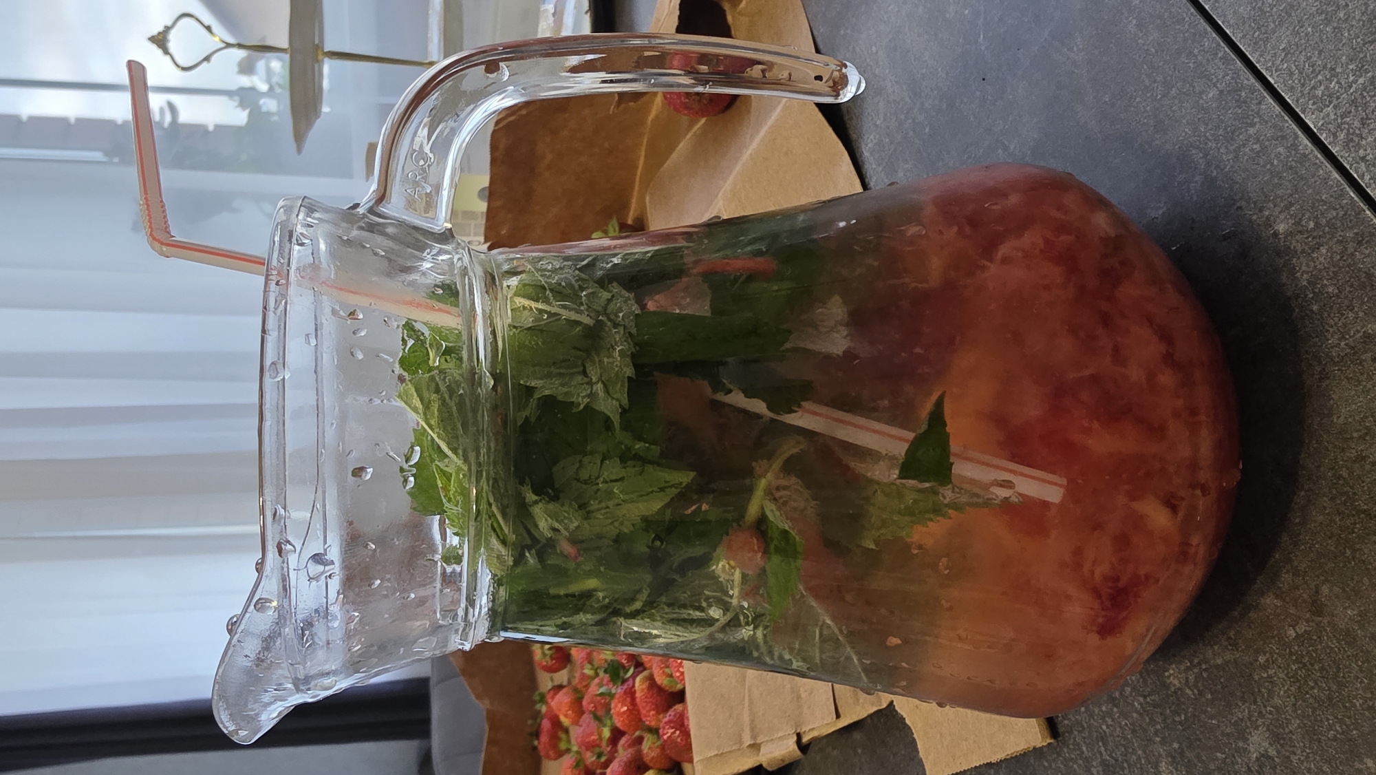 A clear glass pitcher filled with a pinkish liquid, sliced red fruit, and green mint leaves sits on a dark grey tiled surface. Two red and white striped straws are inserted into the pitcher. In the background, a pile of red strawberries is visible on a piece of brown paper. Light filters in from a window with white curtains. A tiered serving stand is also visible in the background. The scene appears to be indoors, possibly in a kitchen or dining area, with a focus on a refreshing beverage. The specific location of Bulgakovo, Russia is not discernible from the visual information provided in the image.