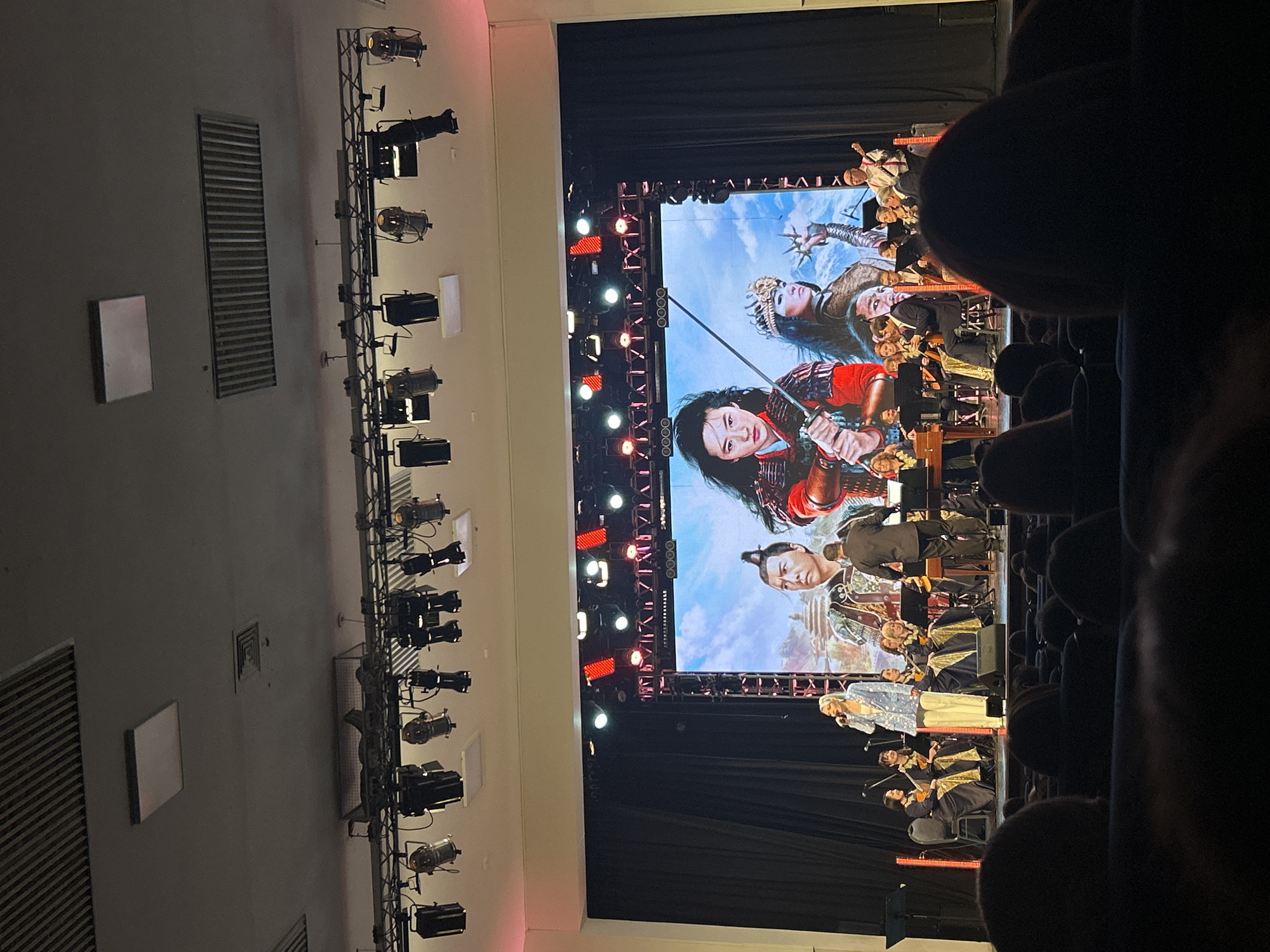 The image shows an audience, likely in a theater, watching a live performance on a stage. A large screen displays images from the movie "Mulan," and a live orchestra and a singer perform on stage in front of the screen. Stage lights illuminate the performance. The location is likely not in Izhevsk, Russia, as "Mulan" is a Disney film, and the image does not contain any visible signs of Russian culture or language.