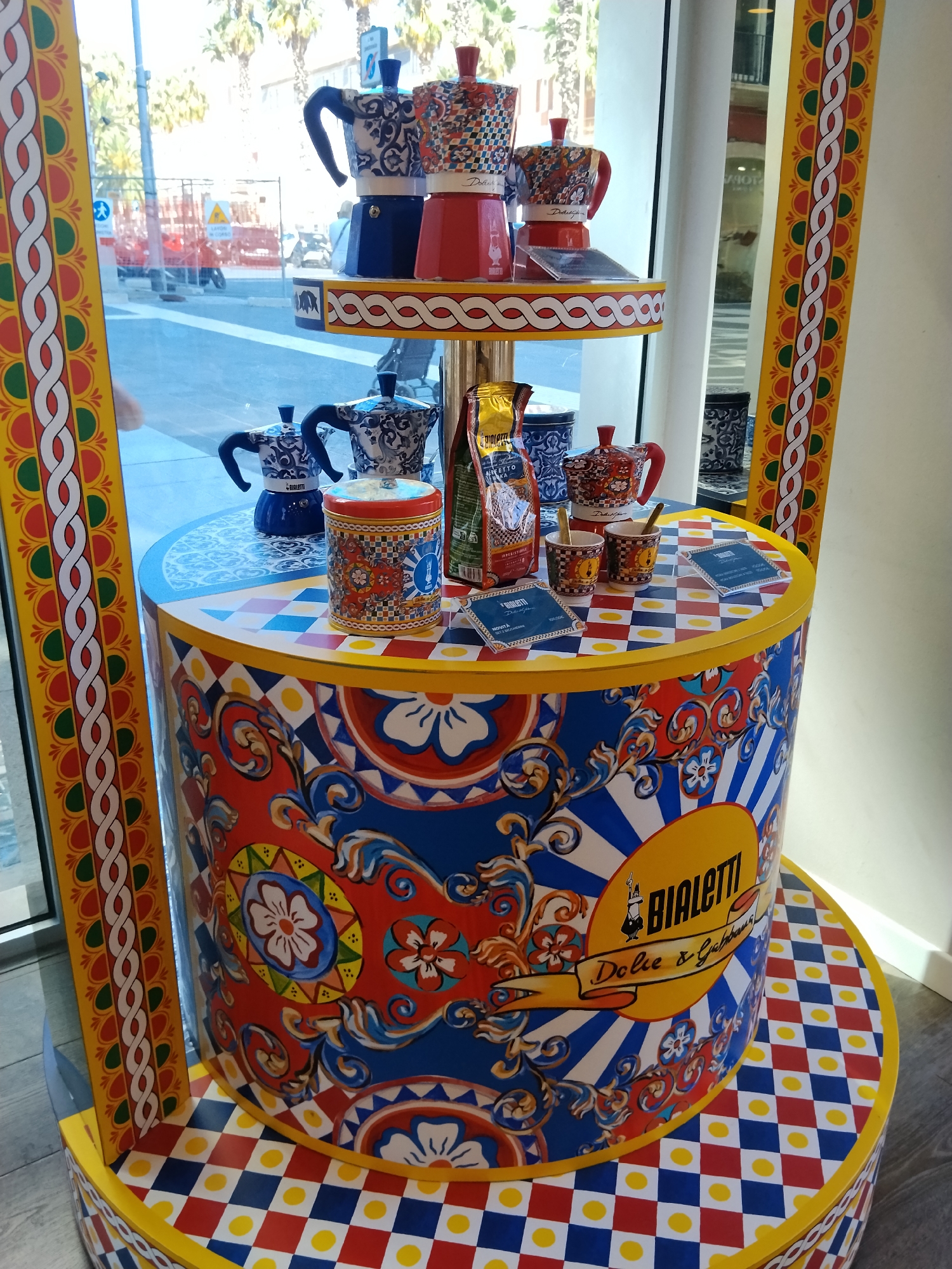 The image shows a colorful display of Bialetti coffee makers and accessories, decorated with the vibrant patterns of Dolce & Gabbana, at a storefront in Bari, Italy. The display is set up in a tiered arrangement. The bottom tier is a large cylindrical pedestal, painted with a detailed floral and abstract pattern in red, blue, white, and yellow. The top of the pedestal features a checkered pattern of red, blue, and yellow squares with yellow circles.
On this pedestal, there are two small coffee cups with spoons, a cylindrical tin decorated with similar patterns to the pedestal, and a bag of coffee. Further up, on a shelf with a red and white wavy border, are three stacked Bialetti coffee makers. The coffee makers themselves are decorated with intricate patterns in blue, white, red, and yellow. Two of these coffee makers are visible in their entirety, while the third is partially obscured by another.
The display is positioned in front of a large window, through which a street scene is visible. The street appears to be under construction, with barriers and scaffolding present. Palm trees and buildings are visible in the background, suggesting a sunny, Mediterranean climate. The display itself is framed by vertical decorative elements on the left and right sides of the image, featuring a repeating pattern of red, green, white, and yellow. The overall impression is a visually striking and artistic presentation of kitchenware.