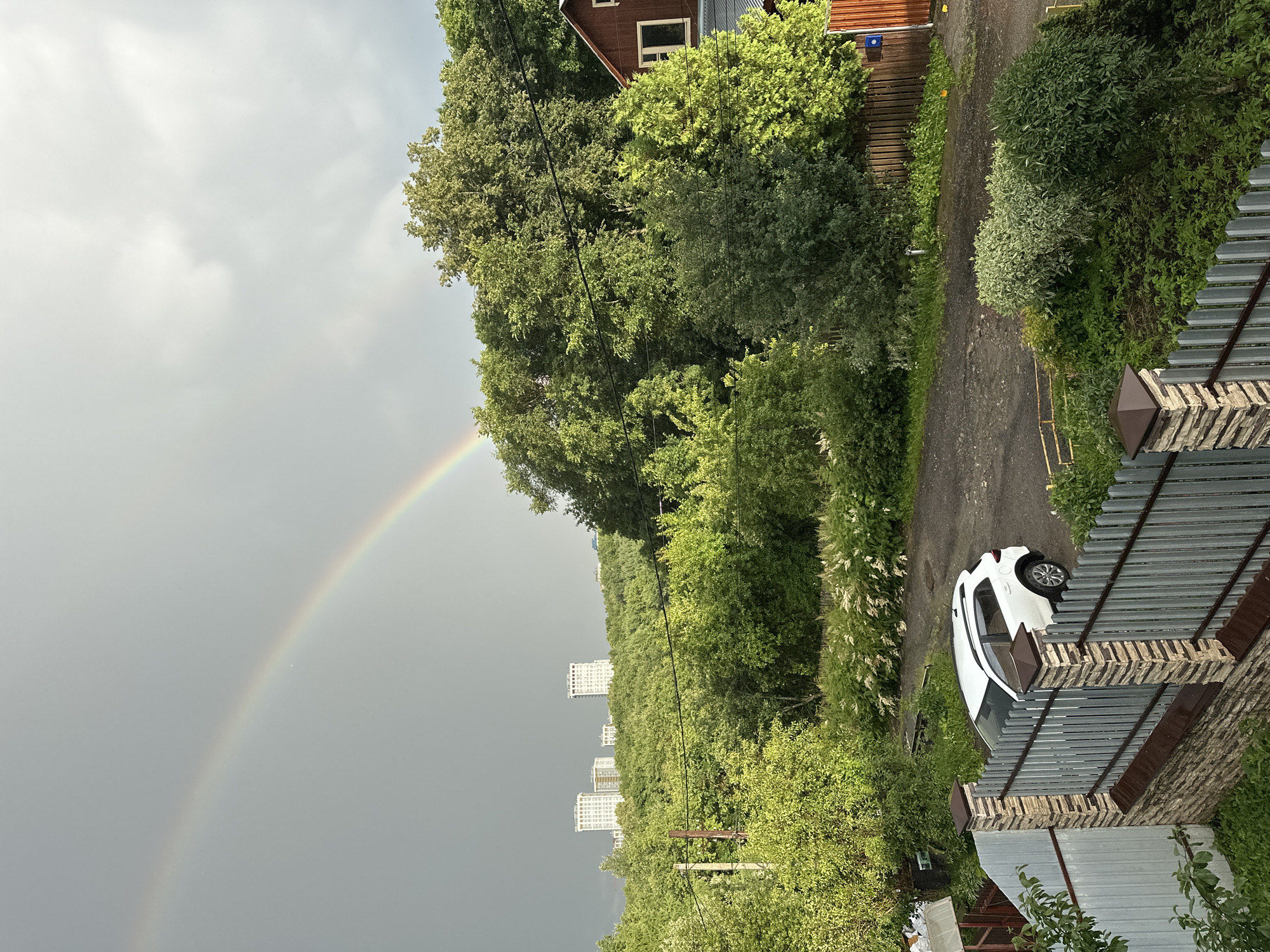 This is a landscape photo taken in Nizhny Novgorod, Russia, on a cloudy day with a rainbow visible in the sky. The scene depicts a residential area with a mix of greenery and buildings.
In the foreground, a white car is parked on a paved path surrounded by lush green trees and bushes. To the right, there's a metal fence and a brick building, suggesting a private property. Further in the background, a dense forest lines the area, and beyond that, several modern apartment buildings are visible, hinting at an urban setting within a natural landscape.
The weather appears to be overcast, with a rainbow arcing across the sky, suggesting recent rain. The lighting is diffused, typical of a cloudy day. The overall impression is one of a suburban or semi-urban environment that experiences natural phenomena like rainbows. There is no visible text in the image.