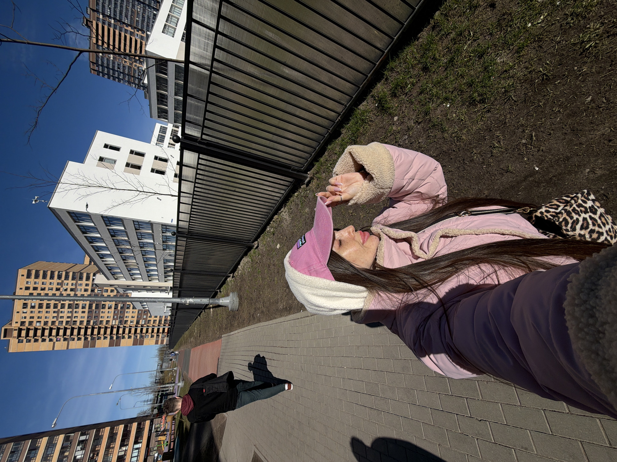 The image shows a young woman taking a selfie outdoors in Saint Petersburg, Russia. She is wearing a pink puffy jacket with a sherpa lining, a pink and white hat, and has a leopard print handbag over her shoulder. She is standing on a brick sidewalk next to a wrought iron fence, with a strip of dirt and grass beyond the fence. Behind her, there are several modern apartment buildings under a clear blue sky. In the distance, another person is walking on the sidewalk. The image appears to be taken with a wide-angle lens.