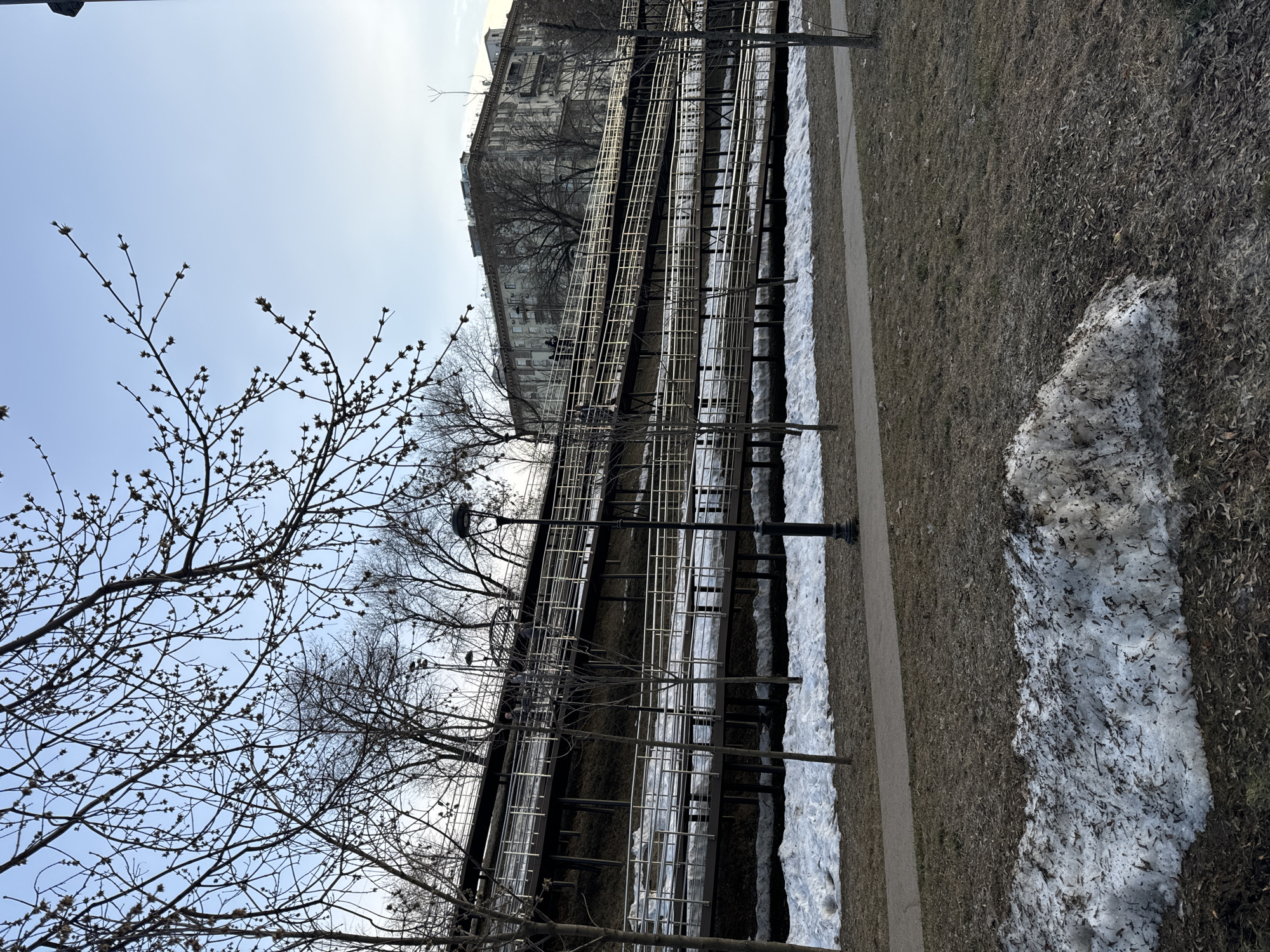 The image displays an outdoor scene in Khimki, Russia, characterized by a multi-tiered pedestrian ramp system ascending a hillside. The ramps feature dark brown platforms and silver-colored metal railings, traversing a brown, sparsely vegetated slope. Patches of melting snow, mixed with dirt and debris, are distributed across the foreground and beneath the lower ramp segments. Dry, light brown grass and scattered fallen leaves cover the ground. A paved pedestrian pathway runs horizontally across the mid-foreground.
Several bare deciduous trees are visible throughout the scene, with their branches prominent against the sky. A black, ornate street lamp stands upright in the mid-right foreground. On the upper segments of the ramp structure, three indistinct human figures are observed walking. In the background, a large, multi-story residential building with a light-colored facade is positioned atop the hill, behind the trees. The sky is clear blue with minimal cloud cover, suggesting ambient daylight conditions.