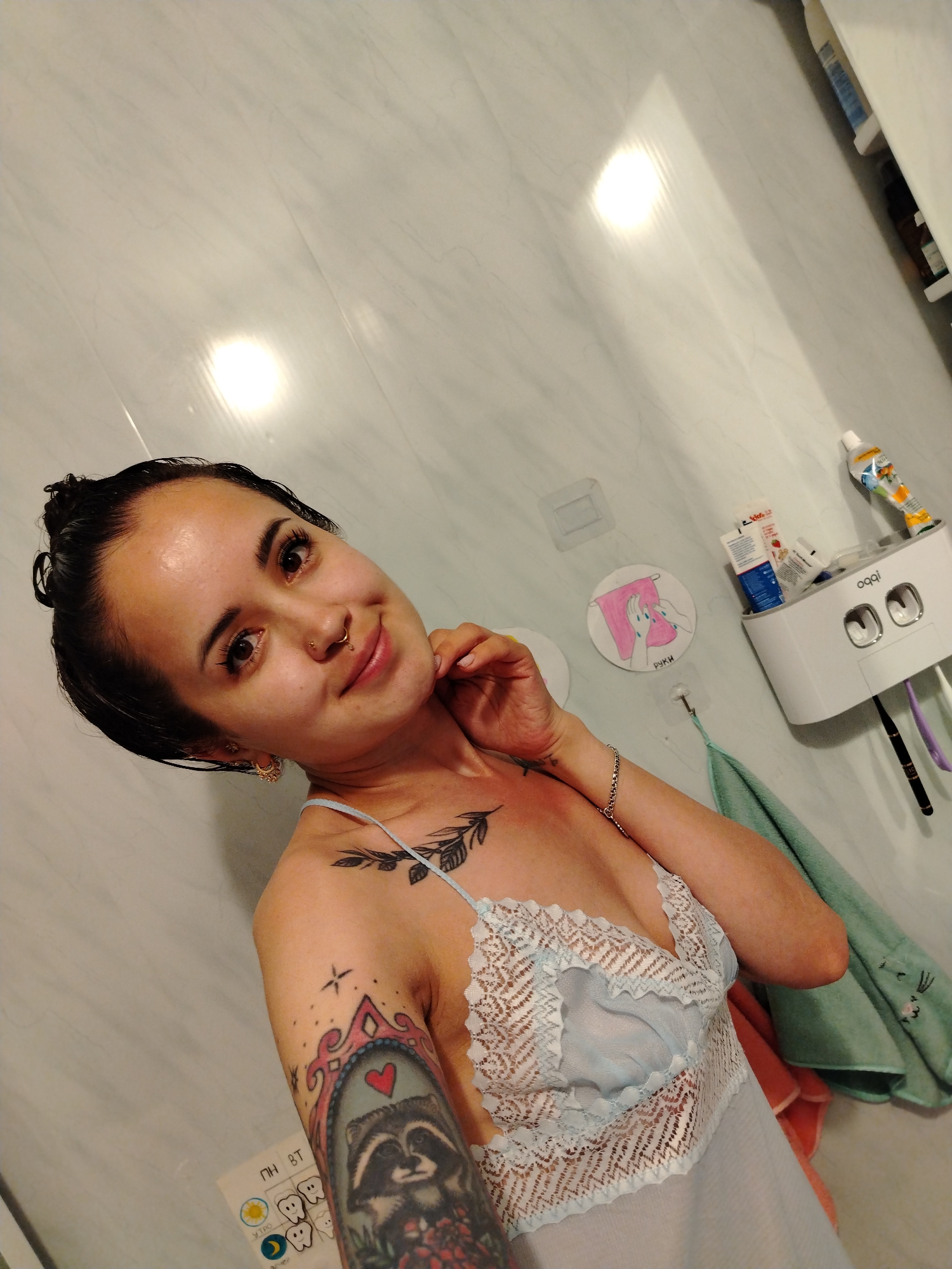 A woman is taking a selfie in a bathroom. She has dark hair pulled back, a nose ring, and earrings. She is wearing a light blue lace nightgown. There are tattoos on her left arm and chest. To her right, a white dispenser for toothpaste and toothbrushes is mounted on the wall, along with two towels. A sticker with a drawing of hands washing is also visible on the wall. The walls appear to be tiled with a marble pattern. The lighting suggests the photo was taken during the day. The location is identified as Noviy, Russia, based on the presence of Russian text on a sticker in the lower left corner.