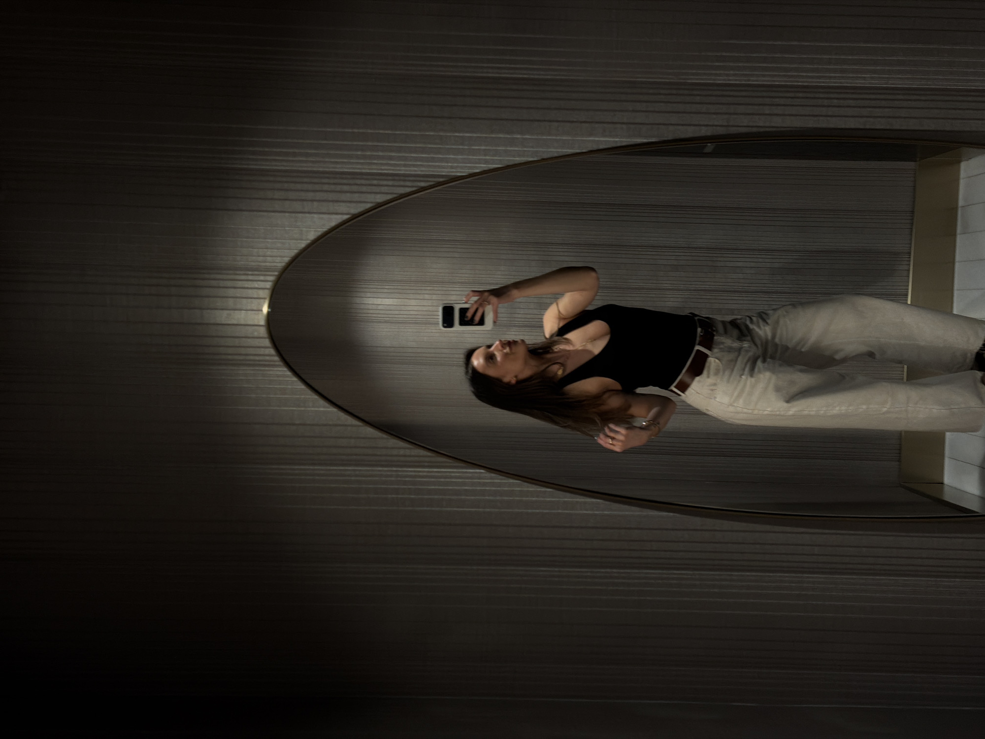 This full-shot image captures a woman taking a mirror selfie in an indoor setting. The photograph is oriented 90 degrees counter-clockwise, showing the woman horizontally within the frame as she stands before a large, oval-shaped mirror. She has long, dark hair and is dressed in a black sleeveless top, light-colored (possibly beige or off-white) wide-leg trousers, and a dark belt. She wears a golden necklace and possibly rings or a bracelet, visible on her right hand which holds a white-cased smartphone, pointed towards herself. Her head is slightly tilted upwards, focused on the phone screen.
The mirror itself has a thin, dark frame and reflects the dimly lit, horizontally textured dark walls of the room. To the right of the mirror, a brighter corner or partition suggests a room division or cubicle. The overall ambiance is sleek and modern, characterized by dark tones and artificial lighting, creating a somewhat intimate or private feel. The setting is likely a stylish hallway, public restroom, or a changing area in a contemporary building, possibly in Budva, Montenegro, as per the location context. There is no visible text in the image.
