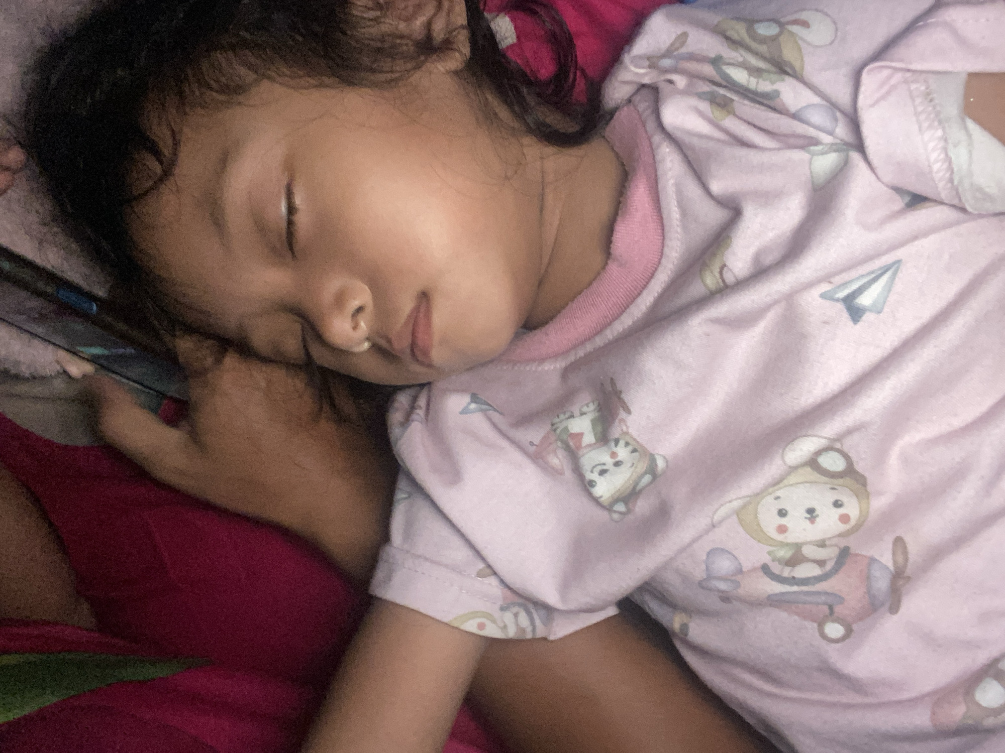 A child is sleeping on a bed. The child is lying on their side, facing the left of the frame. They are wearing a pink top with cartoons printed on it. A hand, presumably belonging to an adult, is holding a phone near the child's head. The bed is covered with a red sheet. The location is Gelam, Indonesia.