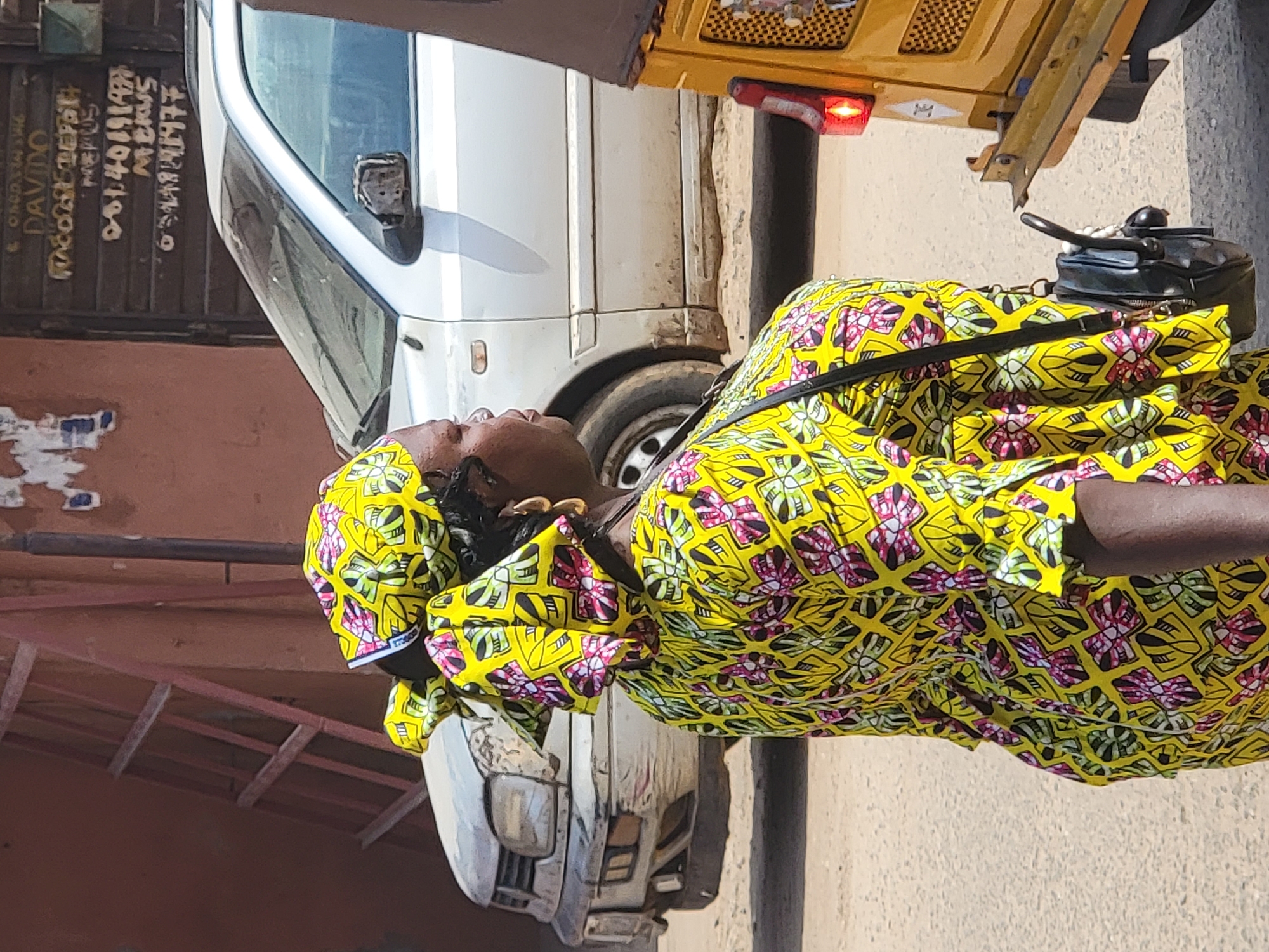 A woman stands on a city street in Maiburiji, Nigeria, wearing a bright yellow dress with pink and green floral patterns and a matching headwrap. She carries a black purse on a strap over her shoulder. Two cars, a white van, and a yellow tuk-tuk are parked near her on the roadside. Text is visible on a nearby structure.