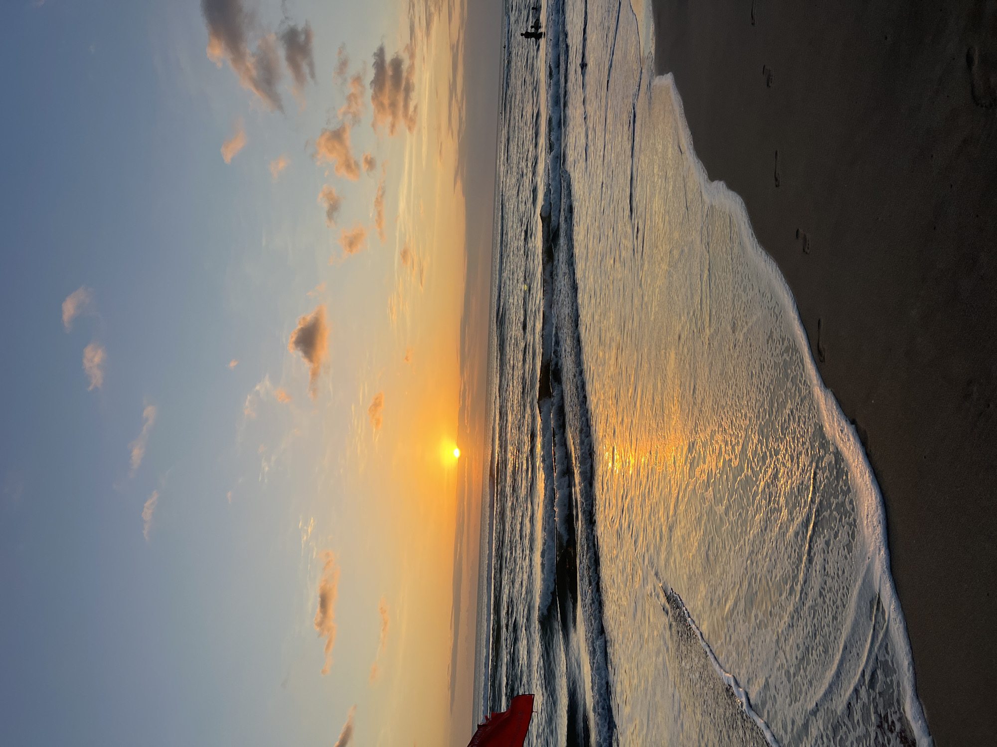 A serene sunset view of Kuta Beach, Bali, Indonesia, is depicted. The sun, a brilliant orb of orange, hovers just above the horizon, casting a golden, shimmering reflection on the gentle waves. The sky is a gradient of soft blues and oranges, dotted with scattered clouds tinged with pink and yellow. The foreground shows the wet sand of the beach, marked with a few footprints, as a foamy white wave recedes. To the left, a torn red flag is partially visible, planted in the sand. In the distance, a few silhouetted figures are seen enjoying the water.