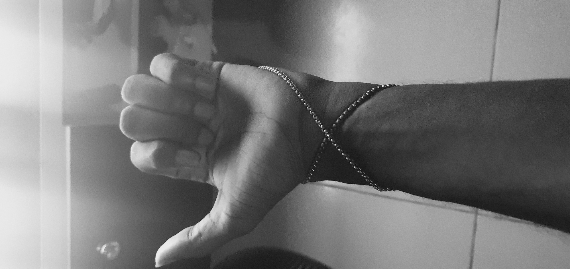 A grayscale image depicts a human arm and hand giving a "thumbs down" gesture. A thin chain is wrapped around the wrist, crossing over the top of the hand. The background includes a blurry image on a wall and tiled wall. The location is not discernible to be Lokoja, Nigeria.