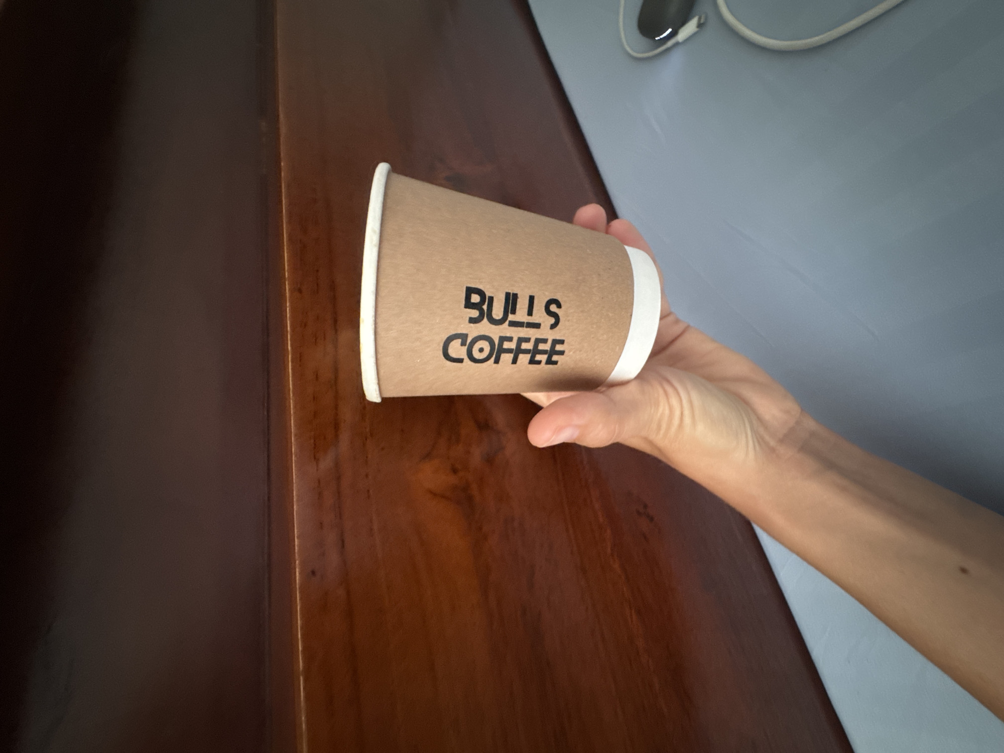 A person's hand is holding a paper coffee cup that has "BULL'S COFFEE" printed vertically on its side. The cup is made of brown cardboard with a white rim. The background consists of a dark wooden surface above a light blue surface, which appears to be a bed or a desk. Some electronic cables are visible on the blue surface. The scene is indoors, and the lighting suggests it is daytime. The location is specified as North Kuta, Indonesia.