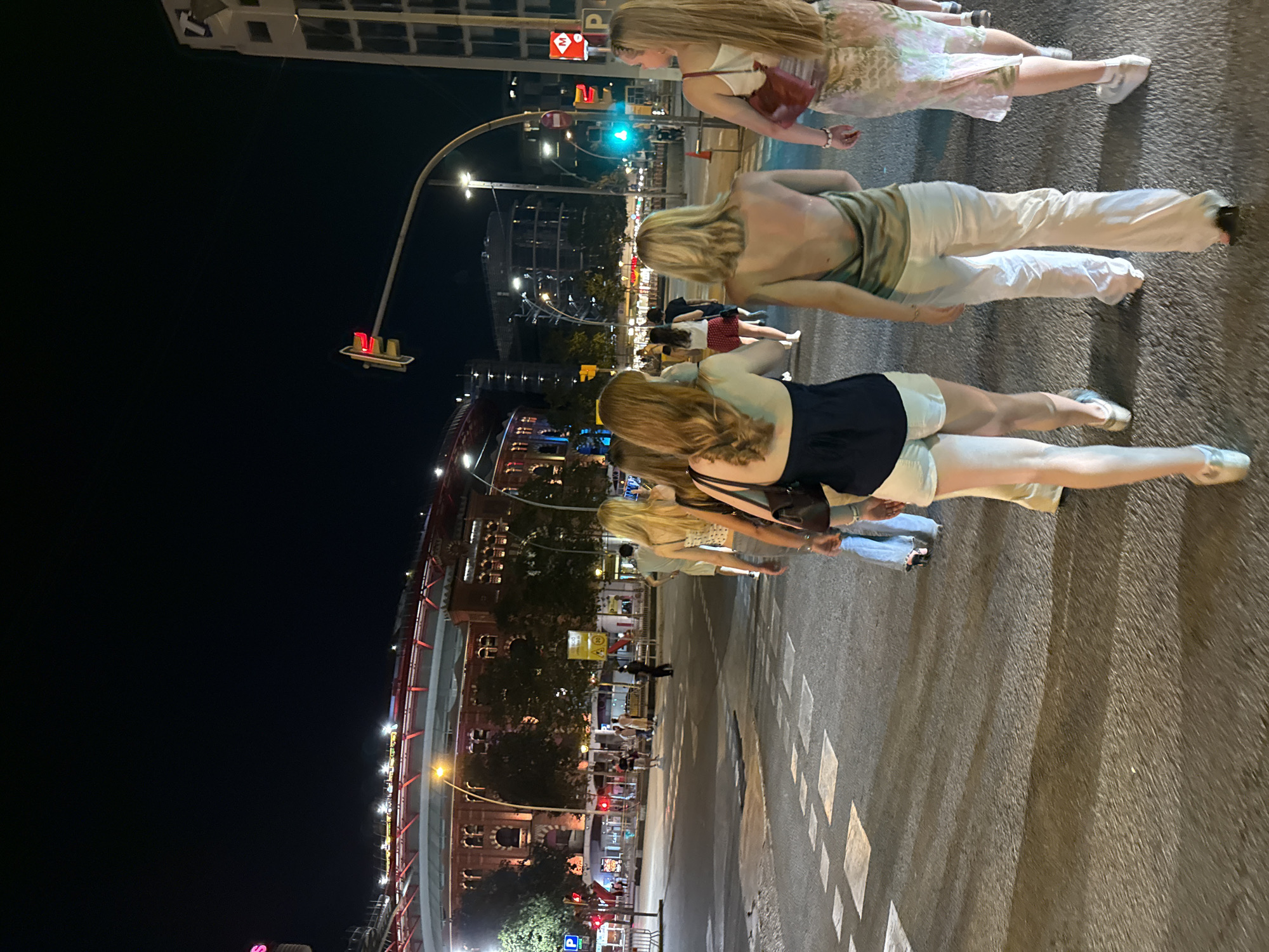 The image depicts a nighttime scene on a street in Barcelona, Spain, near the Las Arenas shopping center, which is visible in the background as a large, circular building with red accents and lights. A group of young women are walking away from the camera towards the left side of the frame, crossing a street. The women are dressed in casual, summery attire. The street is paved, with visible crosswalk markings. Traffic lights are illuminated, with one showing a green light and another showing a red light. Buildings and streetlights are visible in the distance, suggesting an urban environment. The overall atmosphere is one of leisure and social activity.