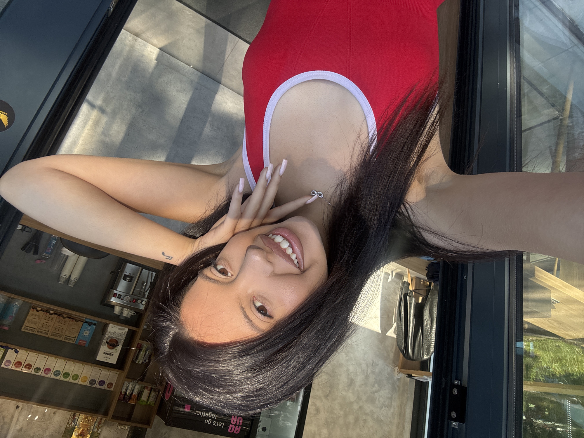 An outdoor selfie captures a young woman with long, dark hair and brown eyes, smiling broadly. She is wearing a red tank top with white trim and an infinity necklace. Her left arm is propped on her head, and her right hand is resting on her chin. The background suggests a café or shop with shelves stocked with coffee products and a coffee machine visible. A glass window reflects sunlight and some greenery, hinting at a location in Sofia, Bulgaria.