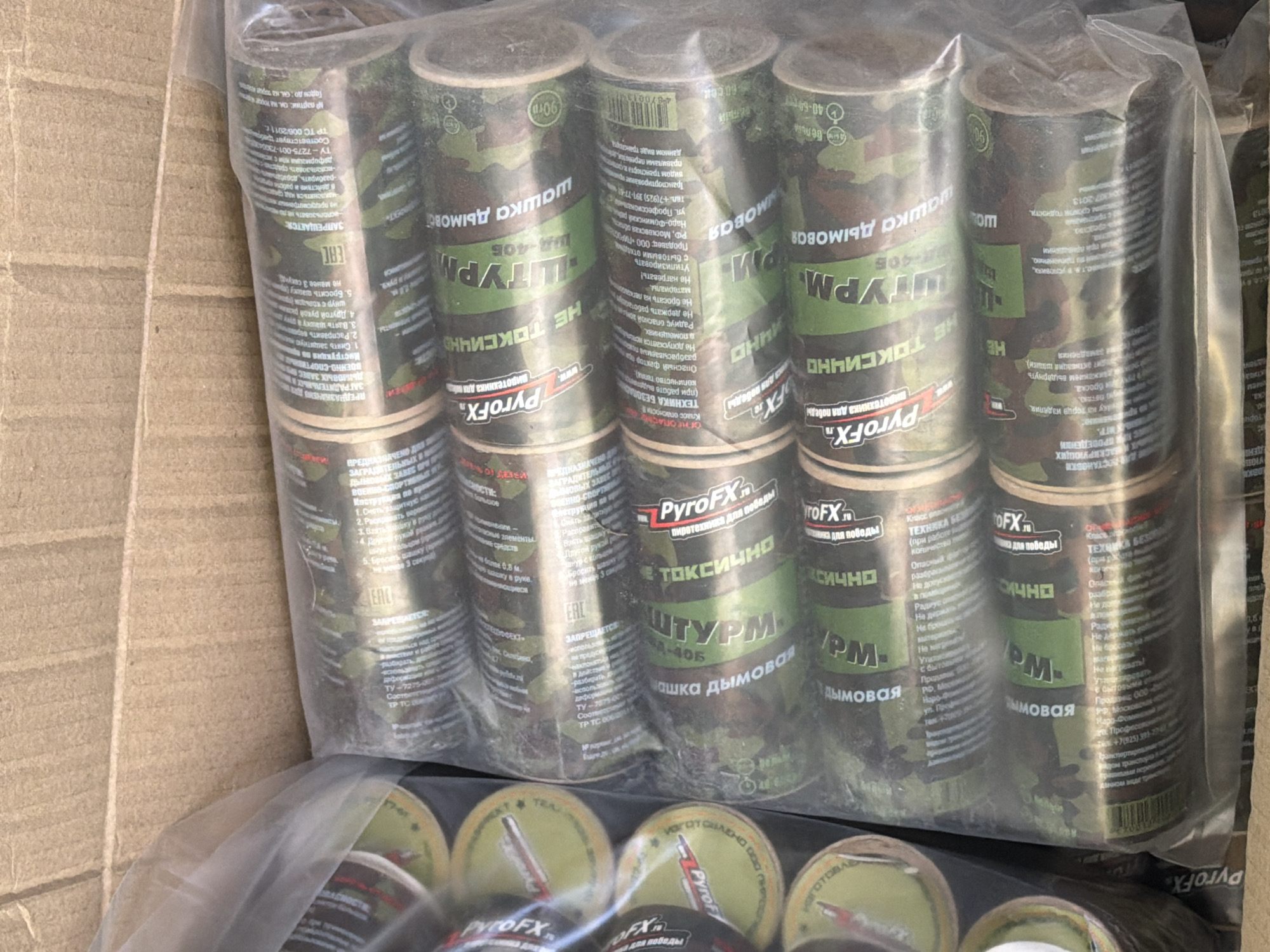 A cardboard box contains multiple cans of "PyroFX" smoke grenades in a clear plastic bag. The grenades are camouflage-patterned and labeled "ШТУРМ ШД-406," which translates to "Shturm SD-406" or "Assault Smoke Grenade model 406." The cans are marked as "НЕ ТОКСИЧНО" (non-toxic). Text on the cans indicates that the manufacturer is located in Naro-Fominsk, Moscow Region, Russia.