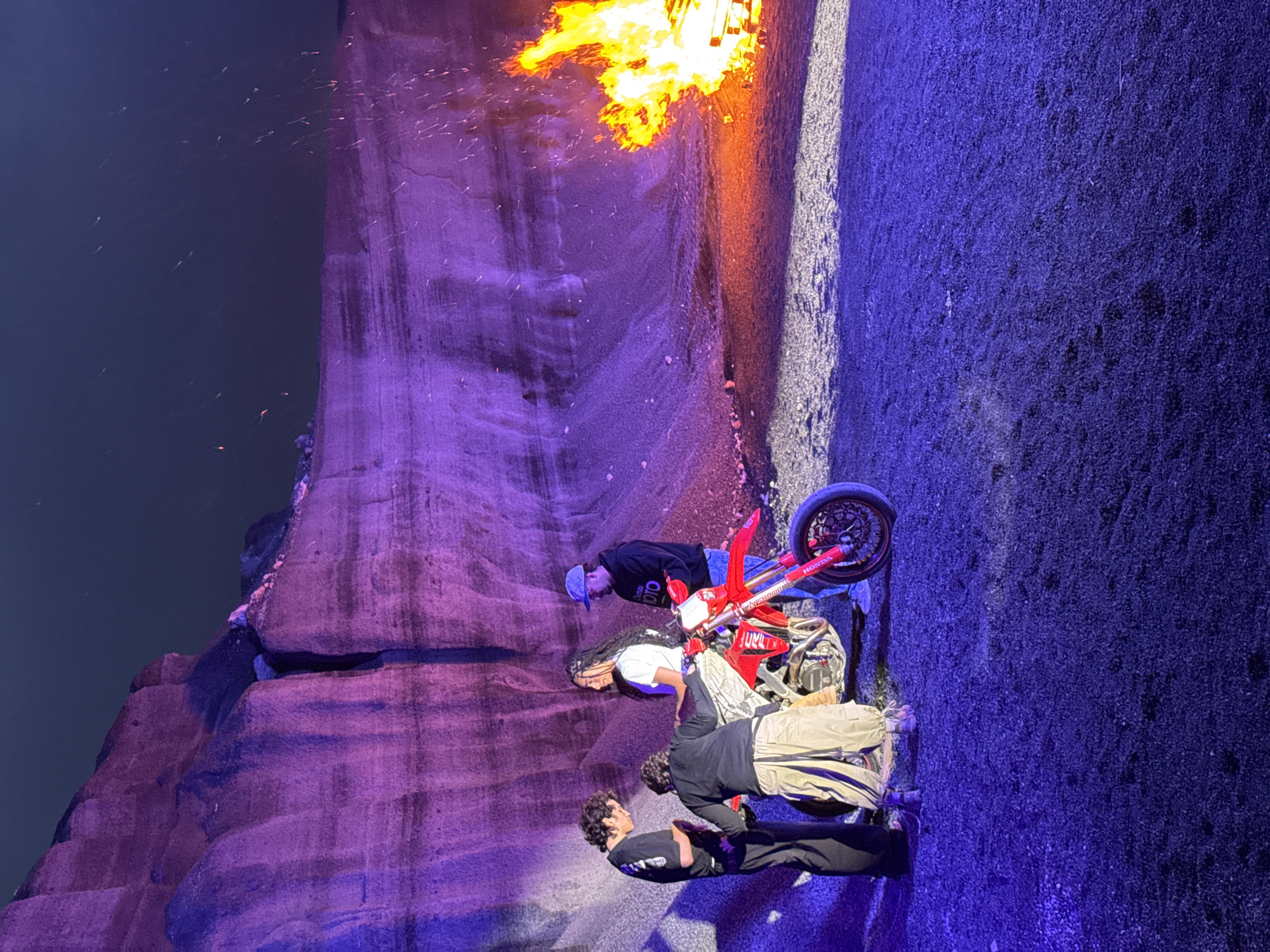 The image captures a scene in what appears to be a performance or event setting, featuring a red Honda motorcycle. It is near a rock formation and a controlled fire. Three people are present. One person is near the motorcycle and faces it, another is on the motorcycle, and the third person is standing nearby. The lighting is dramatic, with strong purple hues illuminating the rocks and blue tones on the ground. The location is likely Teguise, Spain, given the landscape and architectural style.