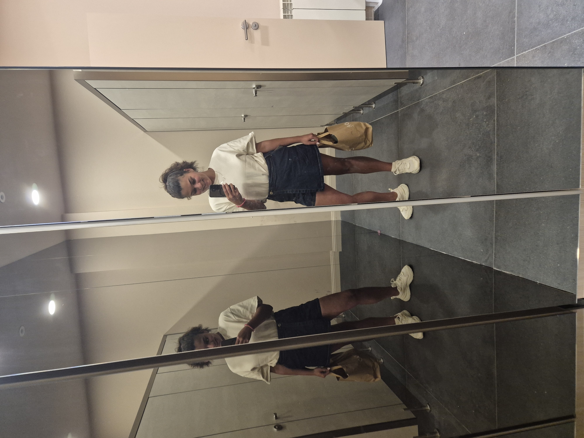 A young woman with dark, curly hair pulled into a ponytail, wearing a loose, cream-colored t-shirt, a dark denim skirt, and white sneakers, takes a mirror selfie in what appears to be a fitting room or changing area. She holds a light brown paper bag in her left hand and her smartphone in her right. The room has light beige walls, dark gray tiled floors, and large mirrored walls, with several changing stall doors visible. The scene is located in Sofia, Bulgaria.