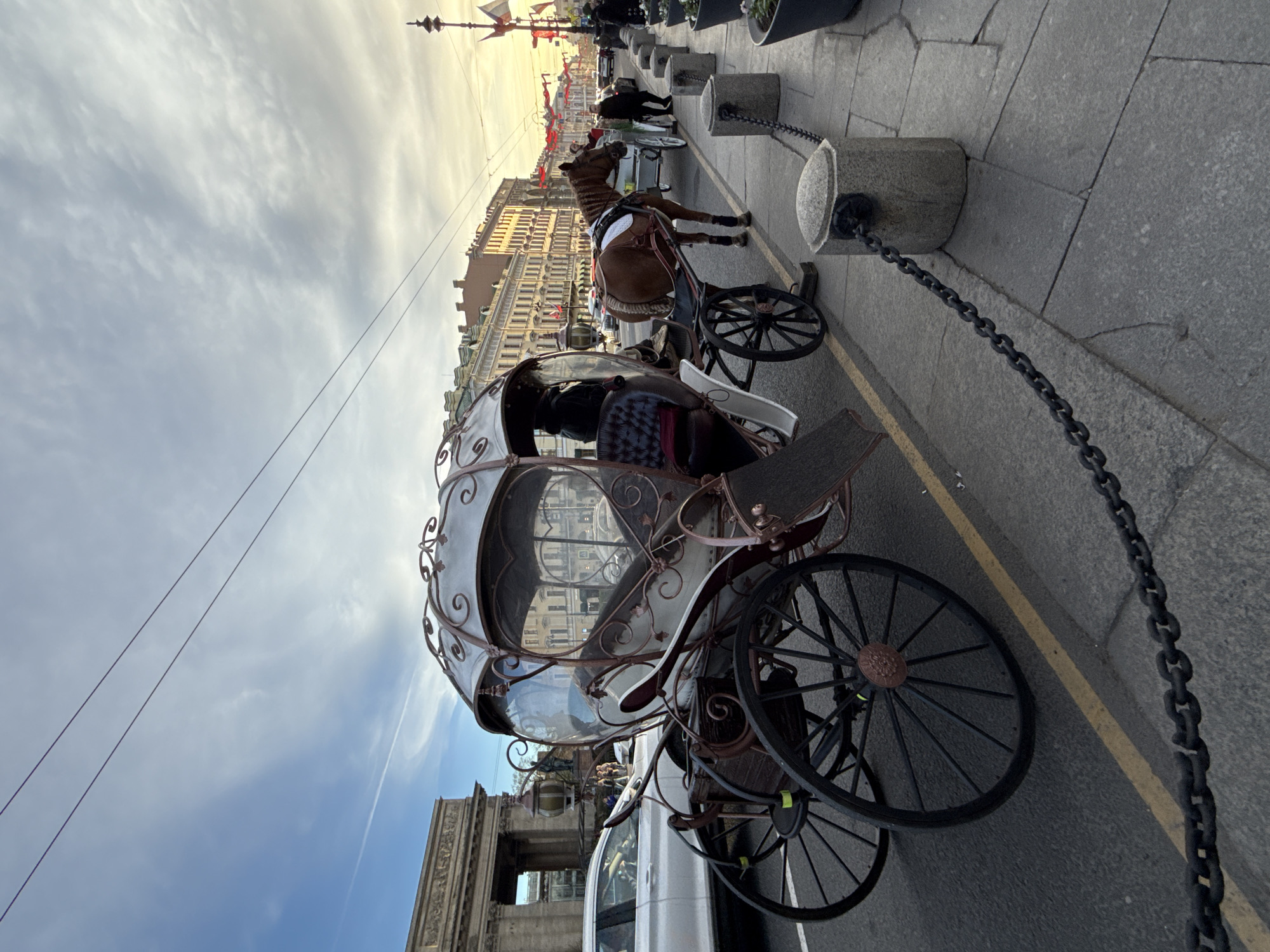 A horse-drawn carriage, likely a phaeton, is prominently featured on a city street, pulled by a brown horse. The carriage is ornate, with a light-colored body, dark spoked wheels, and decorative scrollwork. The horse wears a white saddle pad and harness.
The scene depicts a street in what appears to be a historic European city, confirmed by the context as Saint Petersburg, Russia. The buildings lining the street in the background are multi-story with classical or neoclassical architectural styles, illuminated by the warm, low light of the sun. Small red and white flags or banners are strung across the street between buildings, suggesting a festive or decorative atmosphere.
The carriage and horse appear to be stationary, parked along the curb, possibly waiting for passengers or as a tourist attraction. A dark chain barrier runs along the edge of the sidewalk, connected by stone posts. A yellow line marks the division between the parking area and the road. The sky above is partially cloudy, showing both blue patches and wispy white clouds, indicative of fair weather during the golden hour, either late afternoon or early evening. Overhead wires are visible against the sky. A white car is partially visible parked behind the carriage. No discernible text is visible.