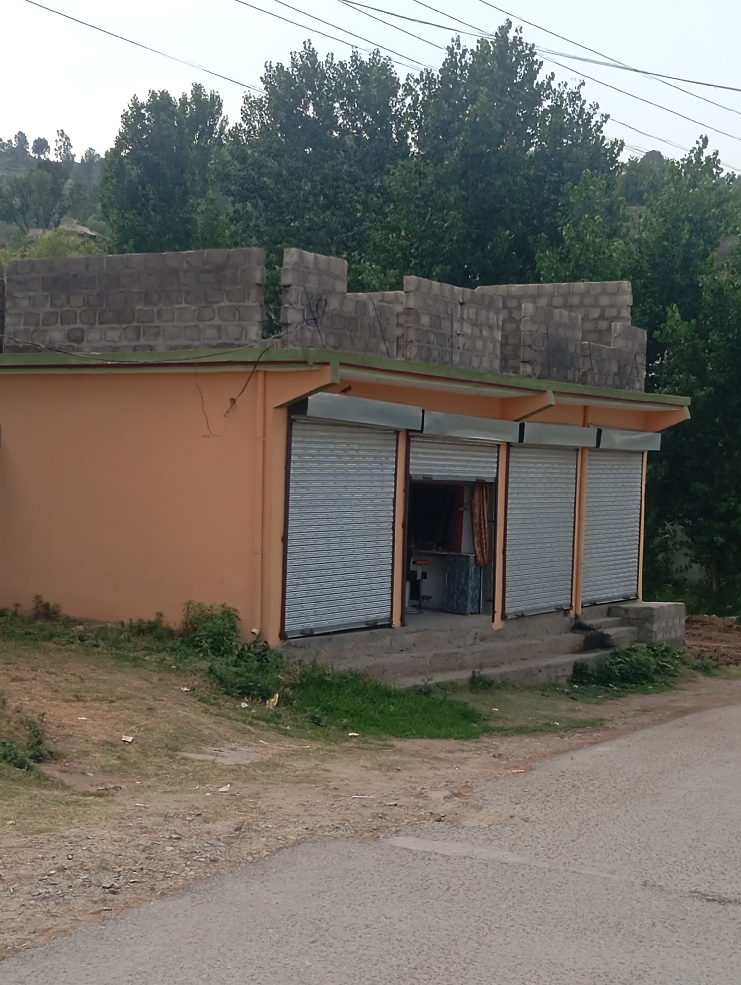 The image depicts a small, single-story commercial building in Lassan, Pakistan. The building has an orange facade and a green trim. It features several rolling metal shutters, some of which are partially open, revealing a small interior with a counter and a stool. The building is situated on a sloped lot with a dirt and gravel foreground, leading to a paved road on the right side of the frame. Behind the building, a dense line of trees forms a backdrop, with some vegetation and partially constructed brick walls visible above the roofline. The sky is overcast, suggesting a diffused light. The scene appears to be a quiet, possibly rural, setting.