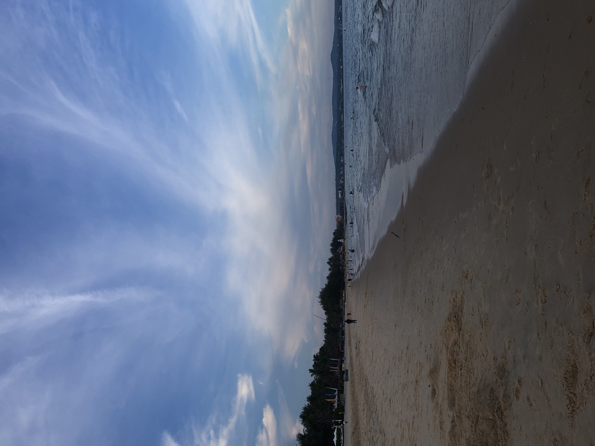This media file depicts a wide-angle view of a beach scene in Phu Thuy Ward, Vietnam. The image is oriented vertically, showing a progression from the sky to the sea and then the sand.
In the sky portion, there are light, wispy clouds against a blue background, suggesting fair weather. The time of day appears to be late afternoon or early evening, given the soft lighting and the way the shadows would likely fall.
The middle section of the image captures the sea meeting the shore. Gentle waves are visible, and there are several people in the water, some appearing to be swimming or wading. A distant shoreline is lined with trees and some structures, hinting at a populated coastal area.
The foreground is dominated by a sandy beach, appearing wet near the water's edge and drier further back. There are footprints in the sand, indicating recent activity. A few scattered objects, possibly debris or natural items, are visible on the sand. One person can be seen walking on the beach.
The overall impression is one of a relaxed day at the beach, with people enjoying the water and the environment. The location cues, such as the type of vegetation and the general appearance of the coast, are consistent with a tropical or subtropical setting. No visible text is present in the image.