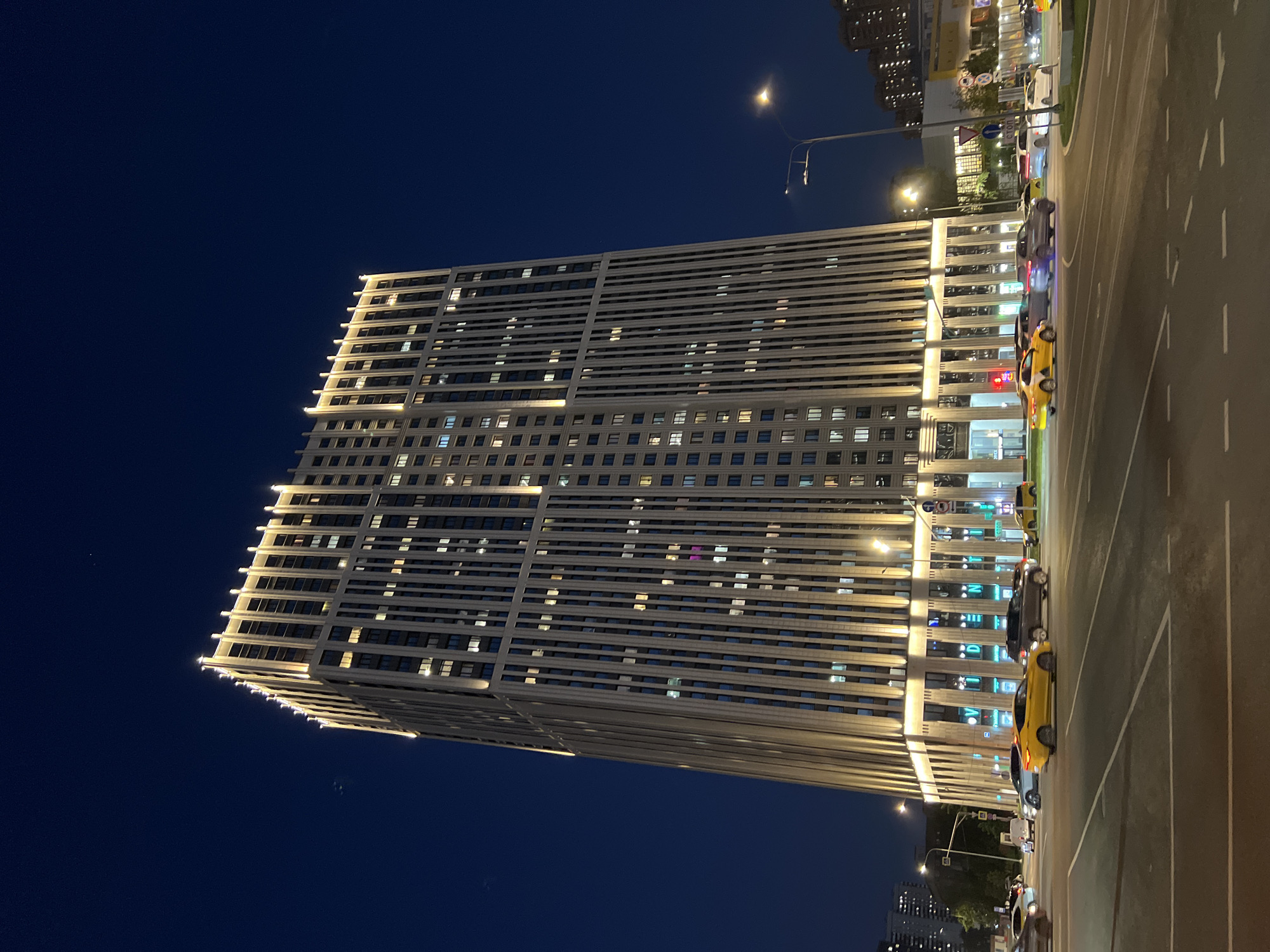 The image shows a tall building at night in an urban environment. The building is rectangular and has multiple stories with many windows. Some of the windows are lit, and there are horizontal lines of lighting along the edges of the building. The sky is dark blue, and streetlights illuminate the scene. A street with multiple lanes runs alongside the building, with several cars parked along the edge of the road. Other buildings are visible in the distance, also illuminated with lights. The location is Unknown, Unknown.