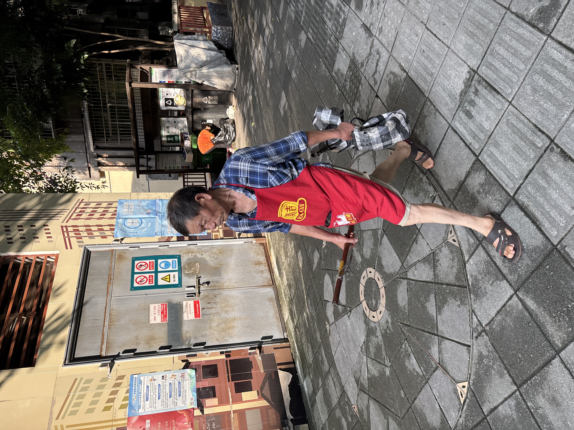 An elderly East Asian man wearing a blue plaid shirt, a red apron, and brown sandals walks on a wet, paved courtyard in Yuzhong District, China. The apron has a yellow emblem with Chinese characters. He is carrying a dark-handled tool in his right hand and a checkered bag in his left. The courtyard is decorated with a circular pattern of stones. Behind the man is a large, weathered metal door with several signs, including a red sign with Chinese characters that reads "Electric Hazard" and a circular green and white sign depicting people in danger from electricity and fire. To the left of the door, a wall is painted with a mural of buildings. To the right, there is a trash sorting area with bins and posters, and beyond that, a residential building. Trees cast shadows on the scene.