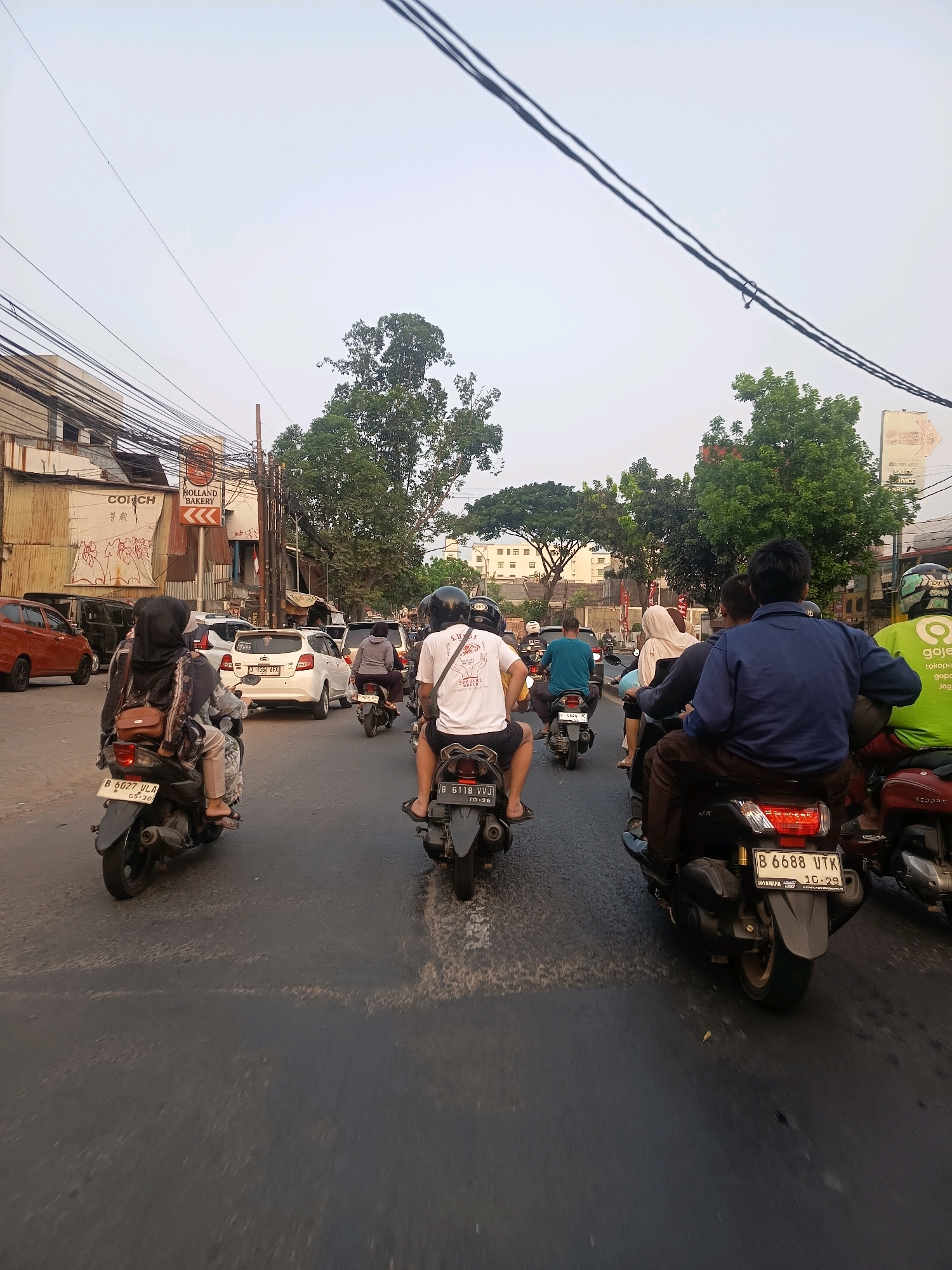 The image depicts a busy street scene in Tangerang, Indonesia, with numerous motorcycles and a few cars navigating the road. The perspective is from a vehicle moving through traffic, facing forward. Several individuals are visible riding motorcycles, some with passengers. The overall atmosphere suggests a typical urban traffic situation in Indonesia. On the left side of the road, buildings and a prominent sign for "HOLLAND BAKERY" are visible, indicating a commercial area. Trees line the sides of the road, and utility wires crisscross overhead, common in many Indonesian cities. The sky is overcast, suggesting either early morning or late afternoon. The road surface is asphalt, with white lane markings. License plates on the vehicles are visible, some with Indonesian alphanumeric characters. The motorcyclists are wearing helmets, a common safety practice. The presence of a Gojek rider, identified by their green jacket, indicates the operation of ride-sharing services in the area.