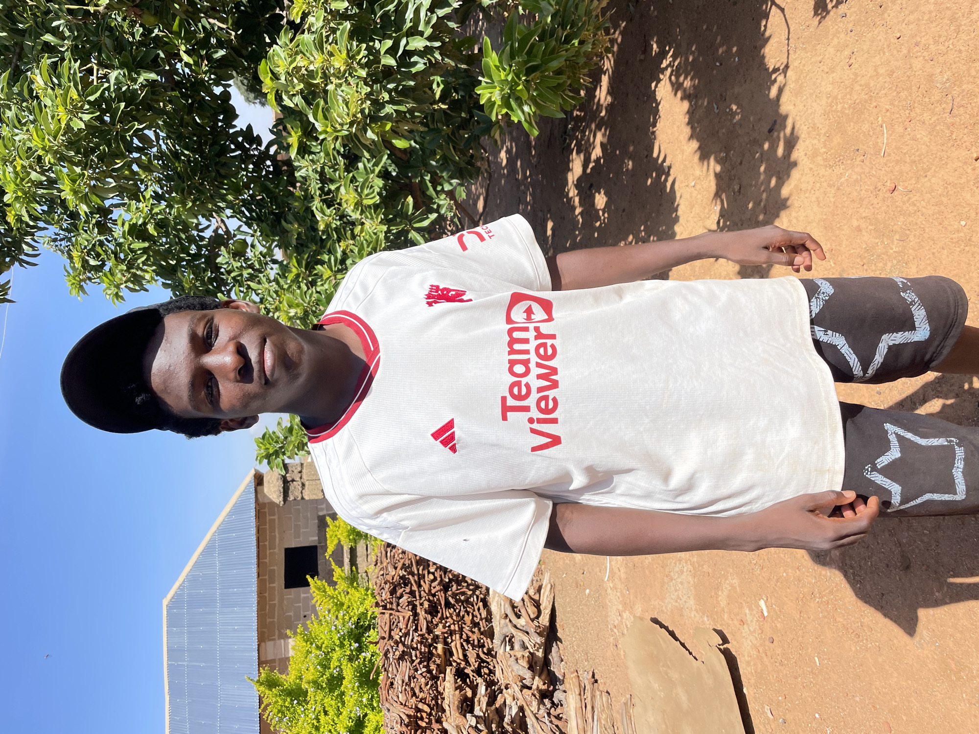 The image presents a young man wearing a white jersey with "Team Viewer" printed in red on the chest, and black shorts with a white star design on the legs, possibly taken in a rural part of Rafin Bauna, Nigeria. The background shows a simple brick building with a corrugated metal roof, a pile of firewood, and green vegetation. The ground is a reddish-brown color, likely soil, and there is a tree providing partial shade. The sky is blue and clear.