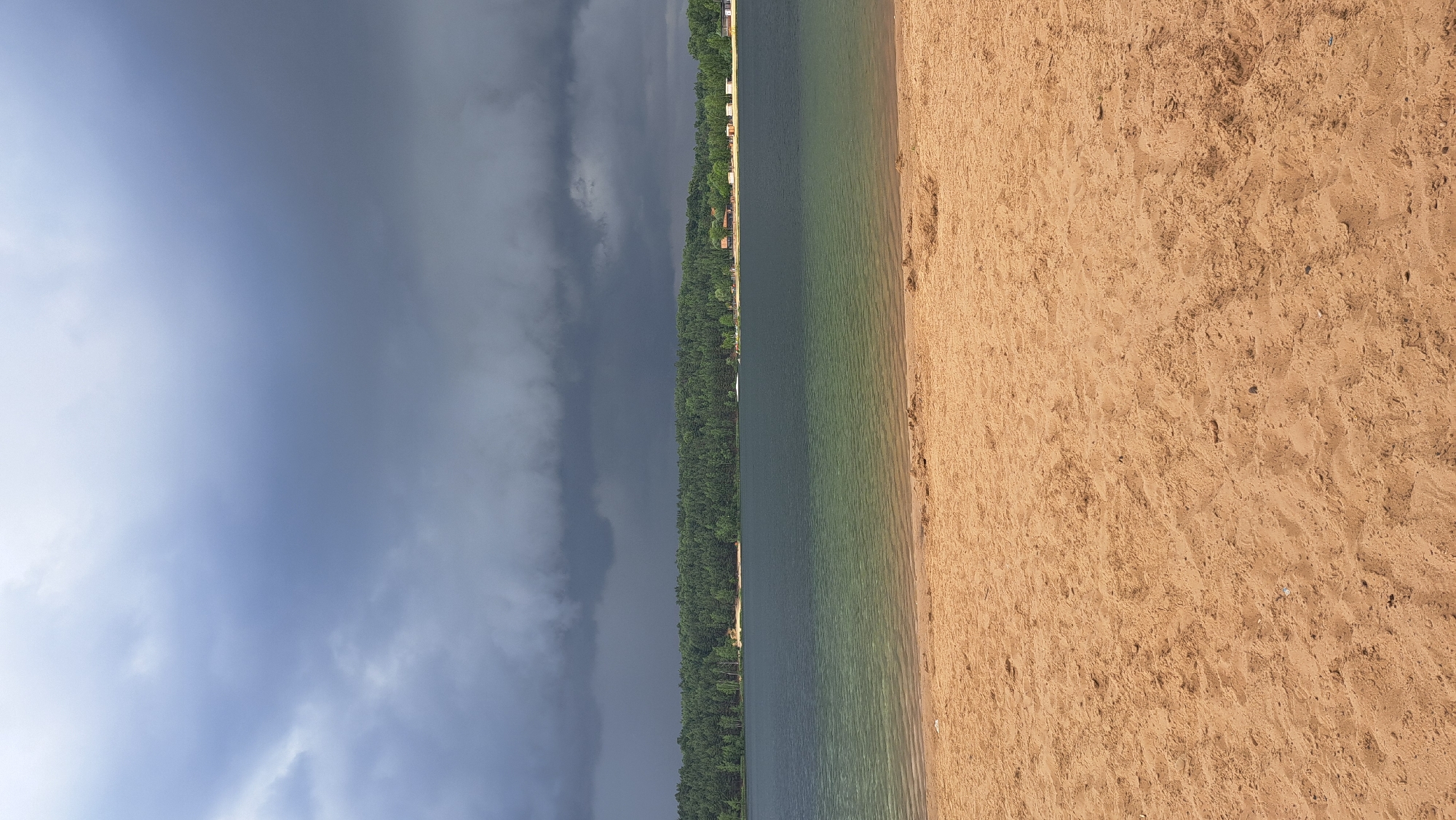 The image shows a wide sandy beach in the foreground, leading to a calm body of water. The water is a deep blue-green, with lighter green and yellow hues closer to the shore, indicating shallow water. A dense, dark green forest lines the opposite bank of the water, with a few scattered buildings visible among the trees. The sky above is dominated by dark, heavy, grey storm clouds, suggesting an approaching or passing weather system. The overall scene depicts a natural landscape at a lake or large pond, likely a recreational area given the presence of a beach and buildings. This location is identified as the city of Lytkarino, Russia, specifically the Lytkarino quarry. There are no people or discernible actions in the image; it is a static landscape photograph.