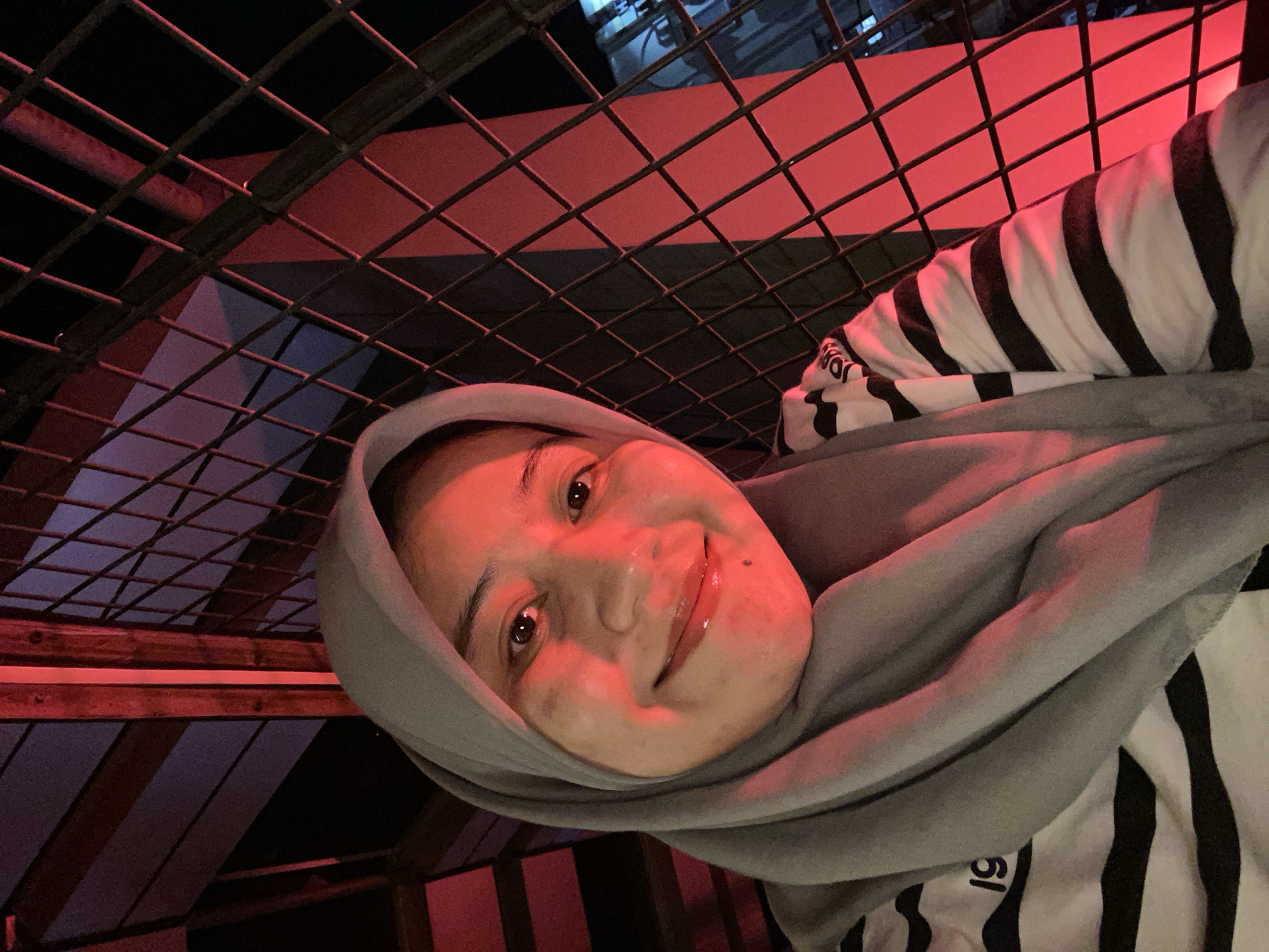 A young woman wearing a grey hijab and a black and white striped shirt is pictured in a selfie. She has light skin, dark eyes, and is smiling. She is standing next to a black metal grate. Behind her, there are wooden beams and what appears to be a building or structure with red lighting. The scene suggests an outdoor setting, possibly at night, with the red lighting creating a dramatic ambiance. The woman's clothing and the overall style of the photograph indicate a casual and personal moment captured by the subject herself. The image does not provide specific clues to pinpoint the exact location within Banjarbaru, Indonesia, beyond a general urban or semi-urban environment.