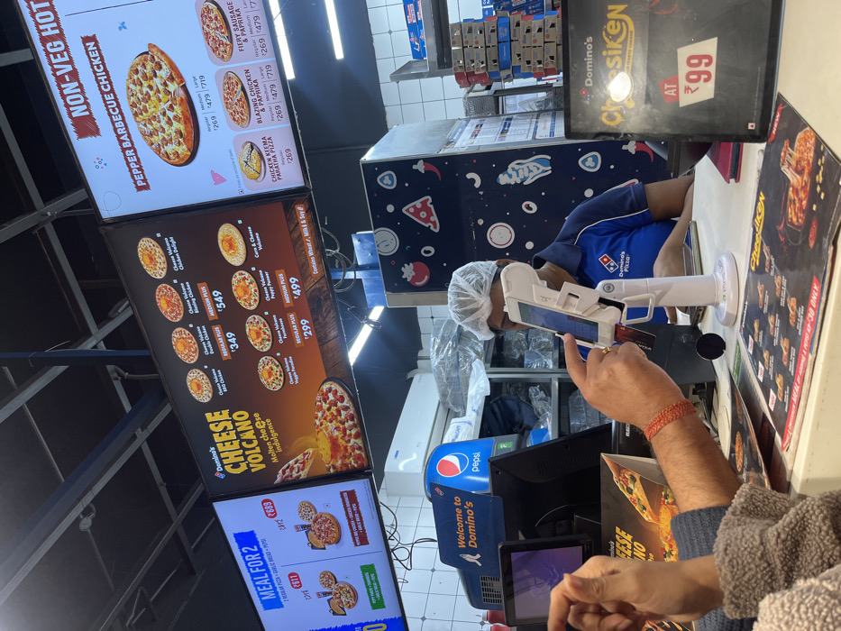 This image captures an indoor scene inside a Domino's Pizza outlet in Tinsukia, India. The setting is a brightly lit counter area where customers place orders.
In the foreground, a customer's arm and hand are visible, engaged in a transaction. The customer wears a red thread bracelet and appears to be holding a card or mobile device near a payment terminal. Behind the counter, a Domino's employee, wearing a blue uniform and a hairnet, is attending to the order, looking down at a screen or device.
Dominating the background are multiple large digital menu boards displaying various pizza options and deals. Visible texts on these screens include "NON-VEG HOT," "PEPPER BARBECUE CHICKEN," "FIERY SAUSAGE & PAPRIKA," "BLAZING CHICKEN & PAPRIKA," and "CHICKEN KEEMA PARATHA PIZZA," with prices ranging from ₹269 to ₹719 depending on size. A prominent section advertises "Domino's CHEESE VOLCANO Molten cheese indulgence," alongside other offers like "REGULAR PLUS ₹349" and "MEDIUM PLUS ₹549," "REGULAR PLUS ₹299," and "MEDIUM PLUS ₹499." One menu board also highlights "MEAL FOR 2." Text stating "Contains Wheat, Milk & Soy" is also visible.
Other notable objects include a "Welcome to Domino's" sign, a Pepsi dispenser, and various stacked boxes behind the counter. A small black sign advertises "Domino's Chosiken AT ₹99," and another mentions "Cheesiken MEAL @ ₹30 ONLY." The overall scene depicts a typical quick-service restaurant transaction.