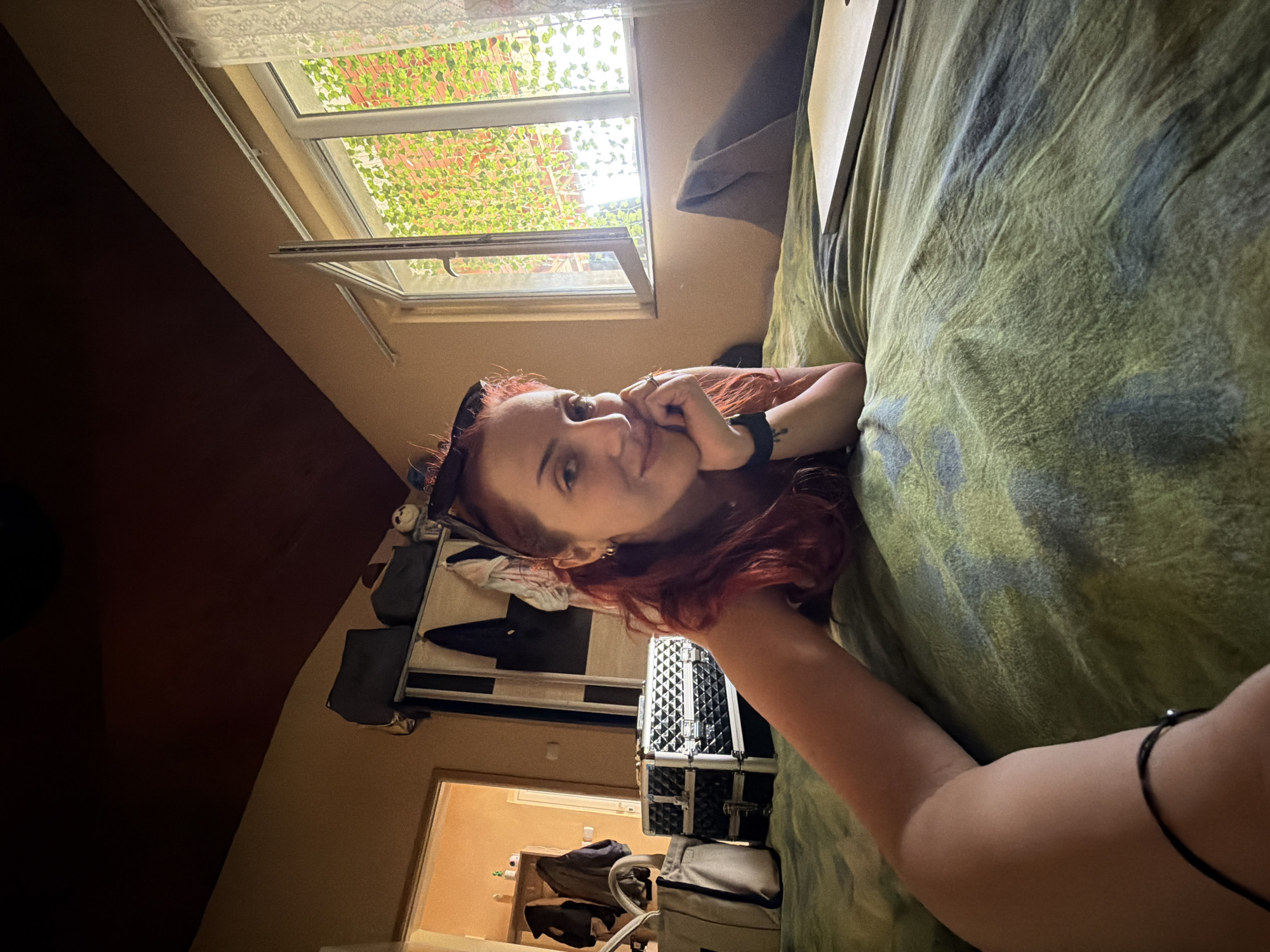 A woman with red hair is lying on a bed covered with a green and blue patterned comforter, looking at the camera and resting her chin on her hand. She wears a black hairband, and there is a black bracelet on her arm. Behind her is an open window with green plants visible outside. In the background, there is a closet and luggage. A doorway leads to another room with objects visible, including a coat and bags.
The image's location is not discernible from the visual information provided. It is impossible to determine if this image was taken in Sofia, Bulgaria, or any other specific location.