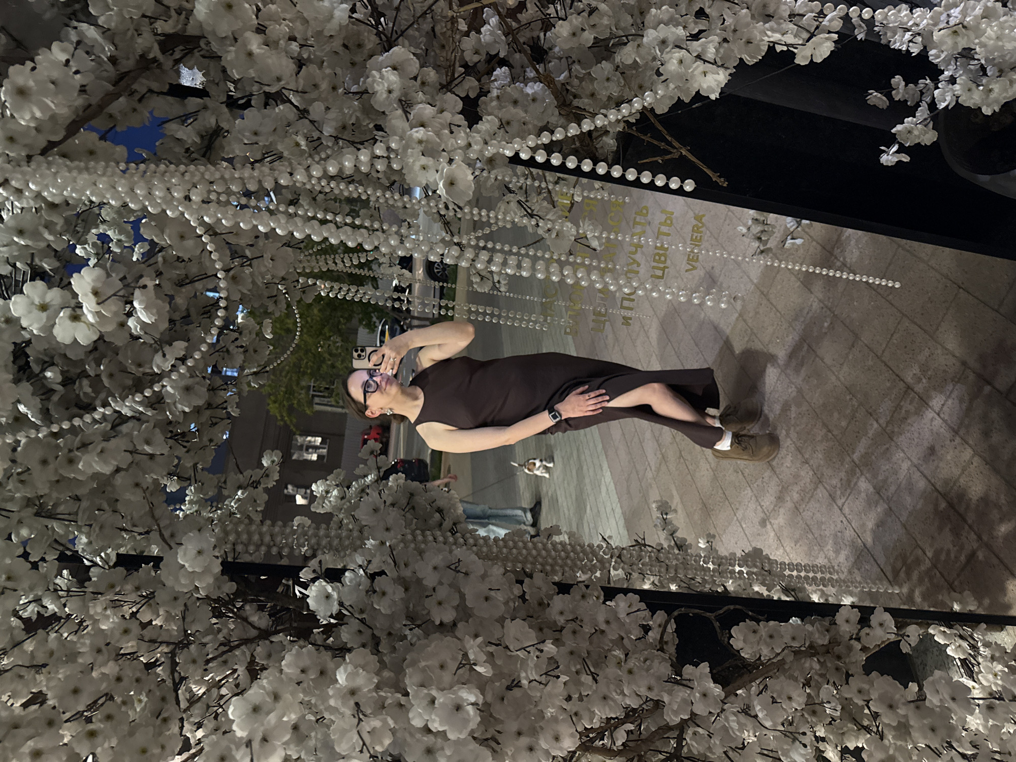 A photo taken at night in Khimki, Russia, features a woman in a brown dress, wearing glasses and a smartwatch, standing near a flower shop adorned with white faux cherry blossoms and strands of faux pearls. She appears to be talking on her phone. A dog and a person are visible in the background. The pavement is made of rectangular slabs. The flower shop has gold text that reads "VENERA ЦВЕТЫ".