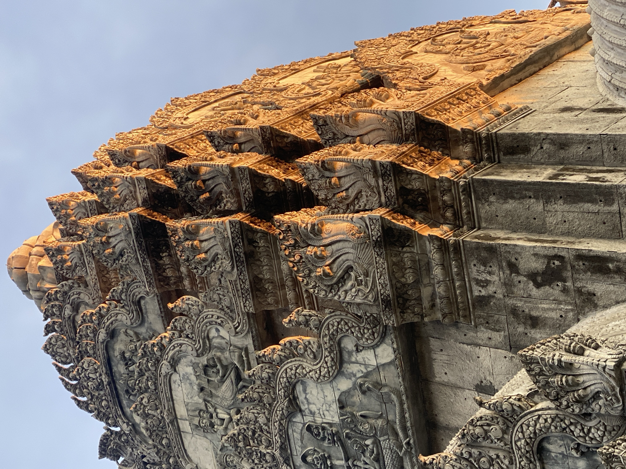 The image shows a detailed, ornate section of a stone structure, likely a temple or similar religious building. The structure is characterized by layered, protruding tiers, each heavily decorated with carvings. The carvings appear to depict mythological figures and intricate patterns, illuminated by a warm, golden light that suggests it may be early morning or late afternoon. The stone is aged and shows signs of weathering. The angle of the shot is looking upward, emphasizing the height and grandeur of the structure against a pale blue sky. This architectural structure is located in Ban Haad Rin, Thailand.