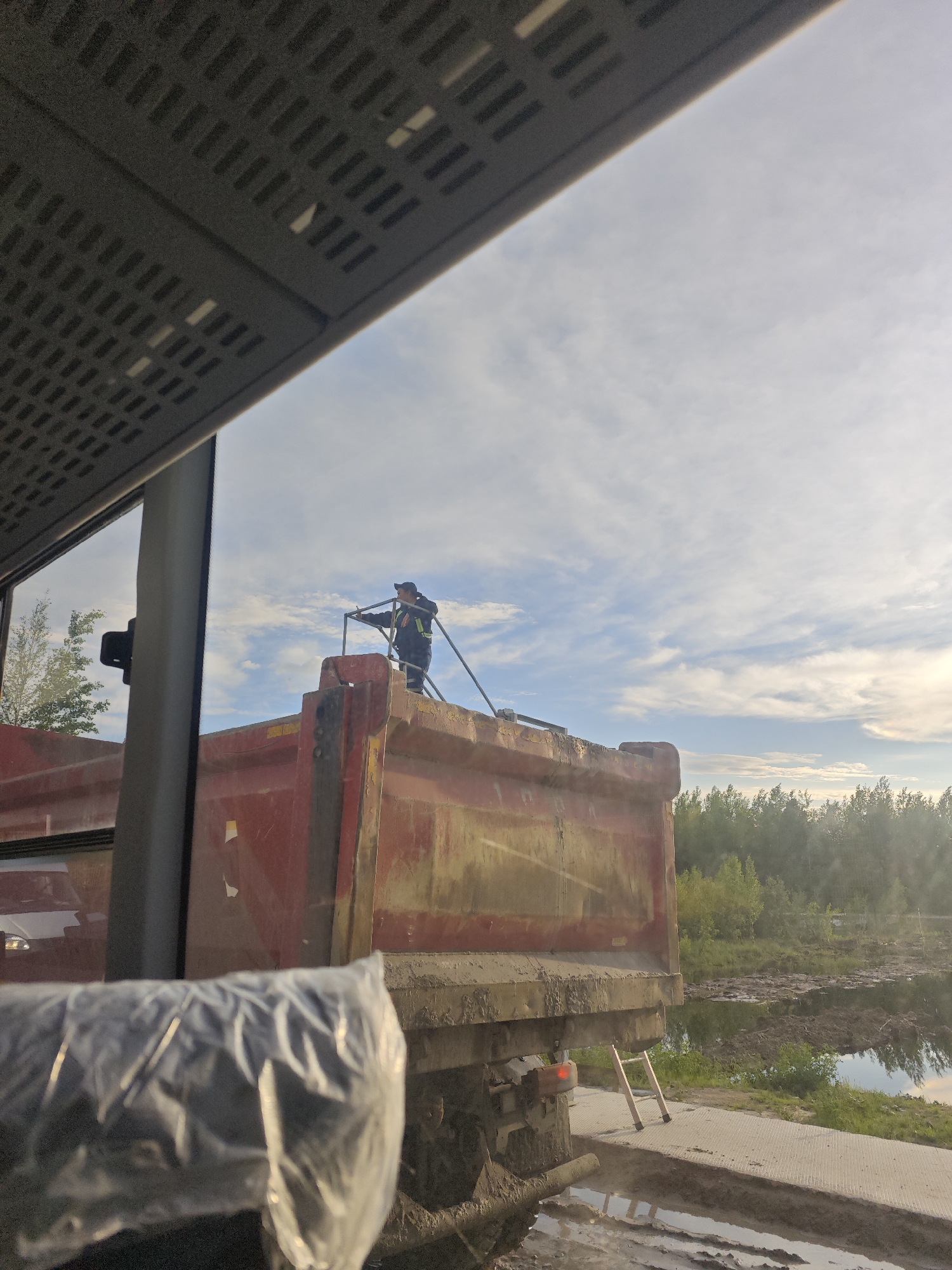 The image depicts a construction or industrial scene taken from inside a vehicle, looking out through a window. The upper left portion of the frame shows the interior perforated ceiling of the observer's vehicle. A large, red, heavily soiled dump truck occupies the central and lower portions of the frame. One individual, dressed in dark work attire with a high-visibility vest and a baseball cap, stands on a metal platform or railing atop the rear of the dump truck. This person holds a long, thin object, possibly a hose or rod, pointed into the truck's bed. Behind the truck, the ground is muddy and uneven, with standing water reflecting the sky. A small, light-colored A-frame ladder leans against the truck's rear frame. To the right of the truck and ladder, a body of water, possibly a large pond or flooded area, reflects the sky and is partially covered with debris or mud. A dense line of green trees forms the distant background. The sky above is partially cloudy with patches of blue. The location is an outdoor work site in the city of Unknown, Unknown.