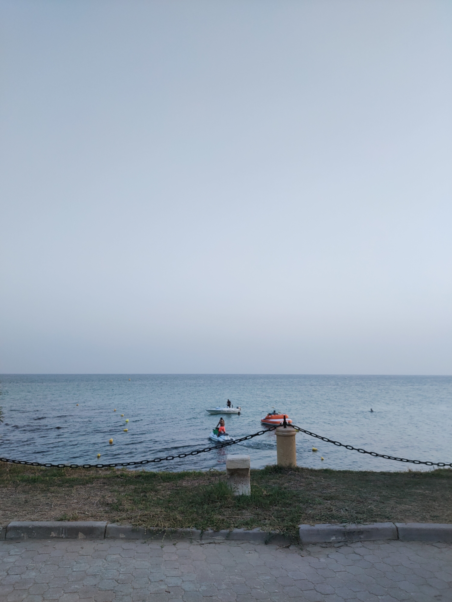 In Nabeul, Tunisia, a serene ocean view is captured with a pale, overcast sky dominating the upper portion of the frame. The calm, blue sea stretches to the horizon, with several small, yellow buoys marking a path on its surface. In the mid-ground, three watercraft are visible: a white boat with two individuals on board, a person riding a jet ski with an orange and white body, and another individual swimming in the water. A black chain fence runs horizontally across the foreground, separating the water from a strip of dry grass and a paved walkway. A stone post with a concrete cap supports one end of the fence. The paved walkway in the very foreground is made of gray interlocking blocks. The overall scene suggests a tranquil day at the beach, possibly early morning or late evening given the muted light.