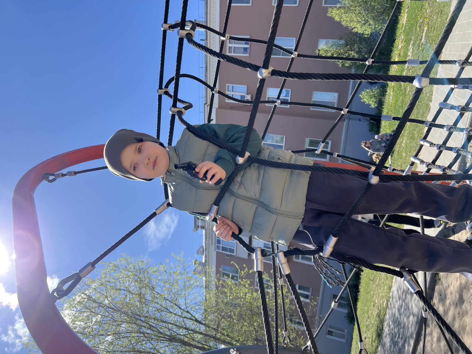 A child is standing on a rope climbing structure at a playground. The child wears a dark hat, light green jacket, and dark pants. In the background, there is a brown apartment building with white-framed windows and greenery on the right. The sky is clear blue. The playground is located in the city of Kazan, Russia.