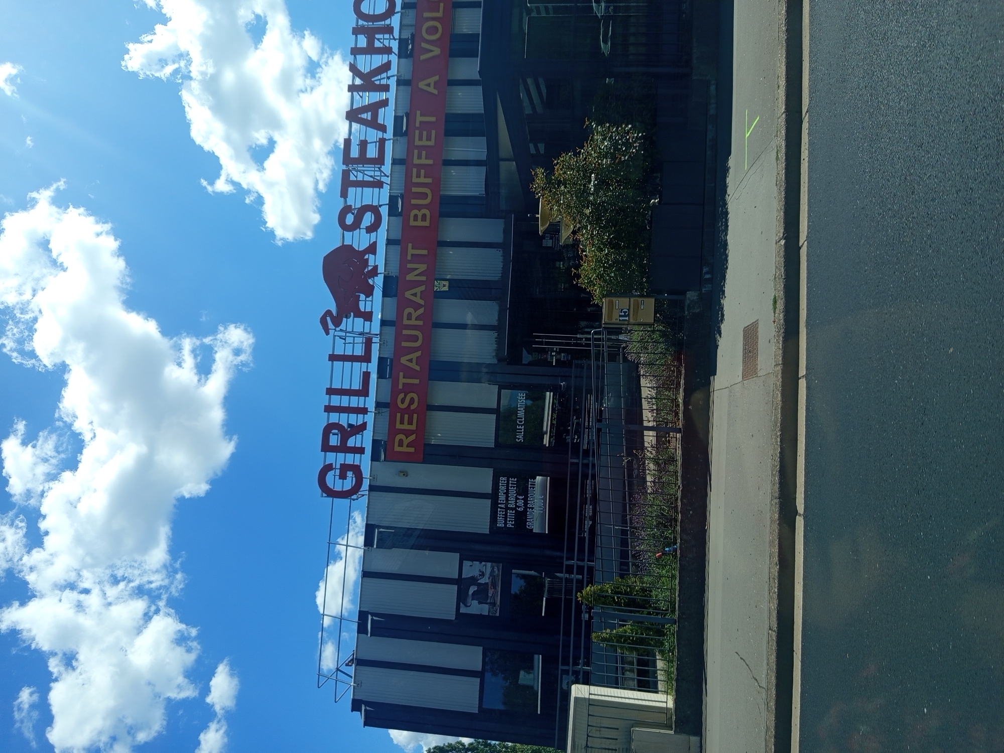 This is a low-angle, outdoor, medium shot of a restaurant with a bright blue sky and white clouds in the background. The restaurant has a large red sign that reads "GRILL STEAKHOUSE RESTAURANT BUFFET A VOL". Below the sign, there are several windows and some greenery. A fence is visible in front of the greenery. A paved road runs alongside the building. The sun appears to be shining, indicated by bright white clouds and the lighting on the building. The image was likely taken during the daytime. Visible text includes "GRILL STEAKHO", "RESTAURANT BUFFET A VOL", "BUFFET A EMPORTER PETITE BARQUETTE 6,00 €", "SALLE CLIMATISÉE", "GRANDE BARQUETTE 11,00 €", and the number "15" on a yellow sign. The setting appears to be a commercial area in Laxou, France. There are no people visible in the image.