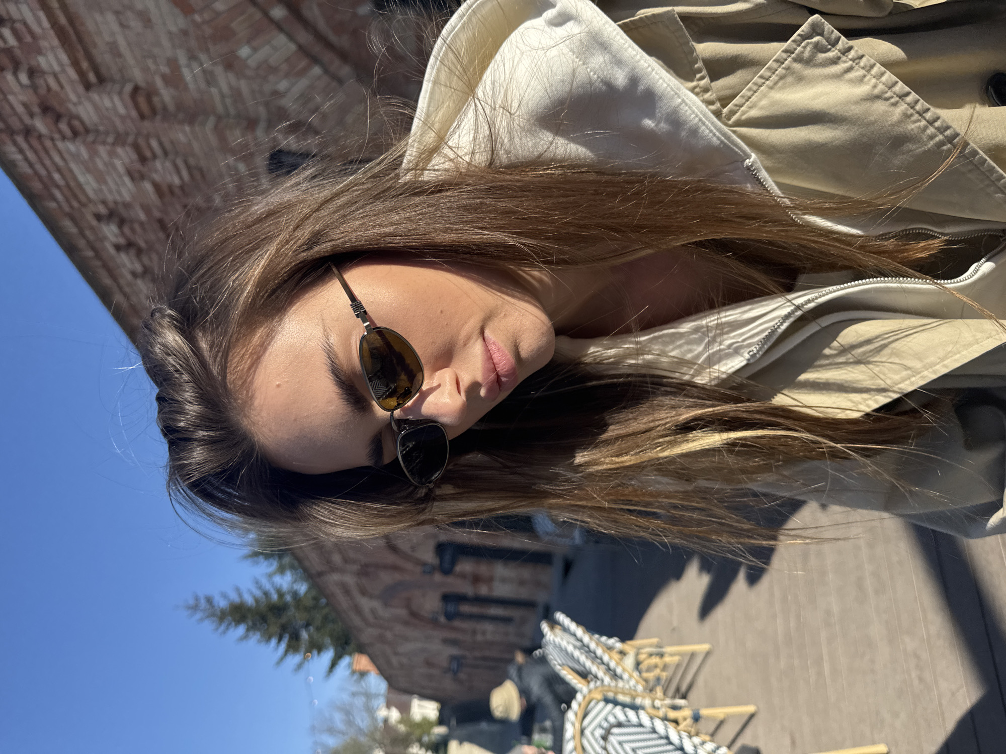 This close-up, angled photograph features a young woman outdoors on a bright, sunny day, likely in Warsaw, Poland. She is positioned with her head tilted, looking downwards, and is wearing dark sunglasses that cover her eyes. Her long, light brown hair falls around her shoulders and against the collar of a light-colored, possibly beige, trench coat or jacket. Her lips are slightly parted, conveying a relaxed demeanor.
The background shows a clear, vivid blue sky dominating the upper left portion of the image. A red-brick building, possibly with dark window features, is visible behind her, suggesting an urban or historic architectural setting. In the lower right, an outdoor patio area is partially visible, furnished with several chairs, some featuring striped or patterned cushions, and what appears to be wooden decking. Distinct shadows are cast across the ground, indicating strong, direct sunlight, suggesting the time of day is mid-morning or afternoon. Scattered green foliage from trees or bushes can also be seen in the distance. The scene evokes a casual outdoor gathering or a moment of leisure on a pleasant day at an outdoor cafe or terrace. No visible text is present in the image.
