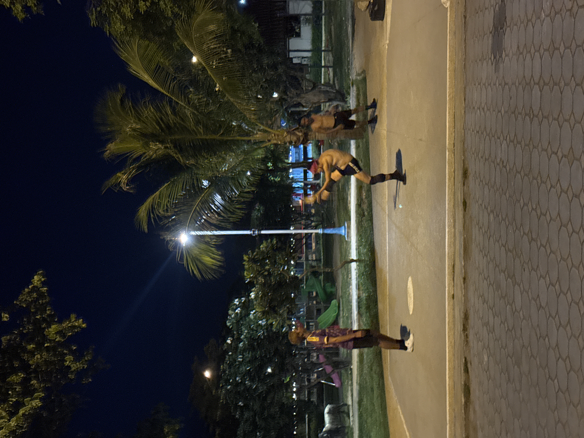 Three individuals are visible on an outdoor court at night in a park in Koh Phangan, Thailand. In the foreground, a person wearing a purple and yellow shirt and dark shorts stands facing left. To their right, a person with pink-dyed hair, wearing black shorts and black knee-high socks, is captured mid-kick, performing a martial arts move. Behind them, another person, also shirtless and wearing dark shorts and black socks, stands observing. The court is bordered by a paved walkway and a grassy area with playground equipment, including a green slide, and trees, including palm trees, are present throughout the scene. A streetlamp illuminates the area.